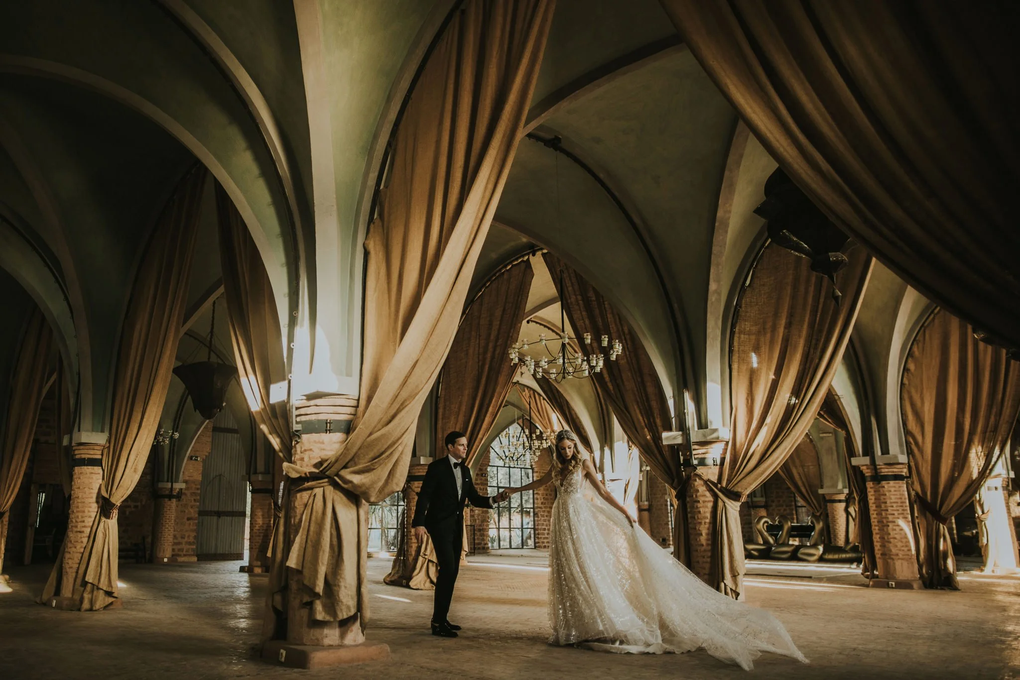 A bride and groom dancing in a grand, arched hall with large curtains, chandeliers, and brick columns at The Beldi Country Club in Marrakech.