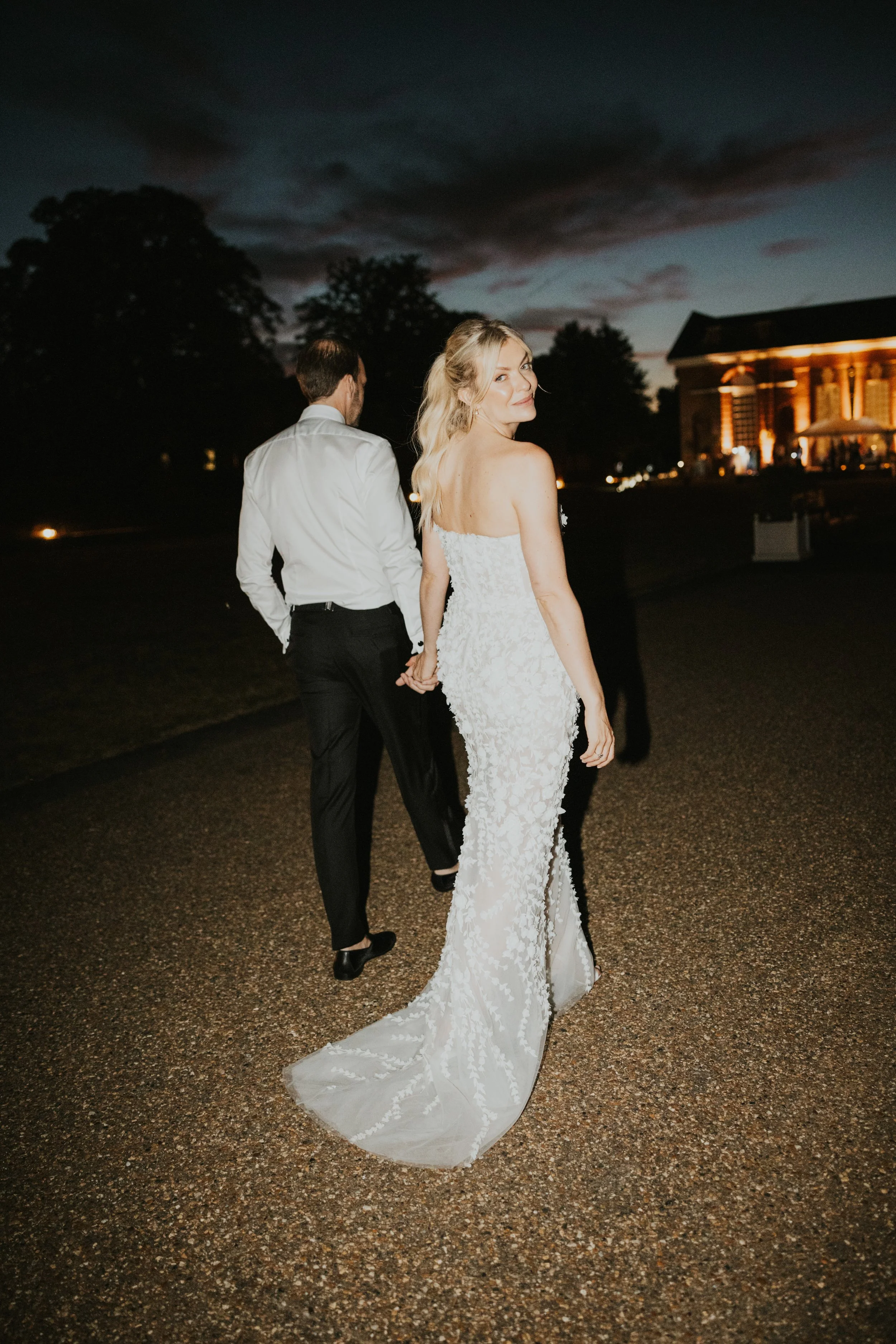Bride and groom Mimi and Simon walking hand in hand at night outside Kensington Palace with the illuminated palace facade glowing behind them