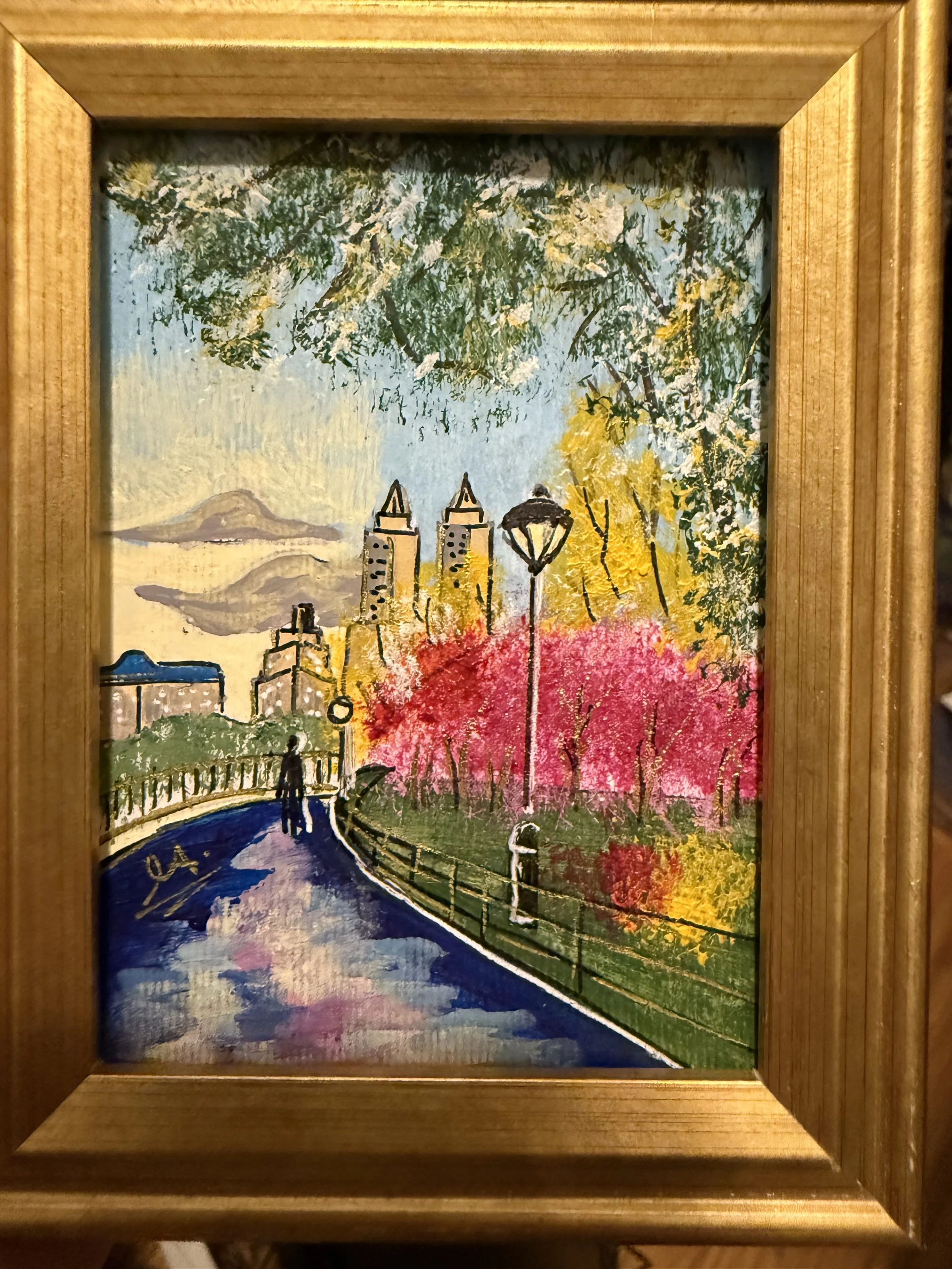 A colorful painting of a city park with a pathway, trees with pink, yellow, and green leaves, street lamps, a person walking with an umbrella, and tall buildings in the background.