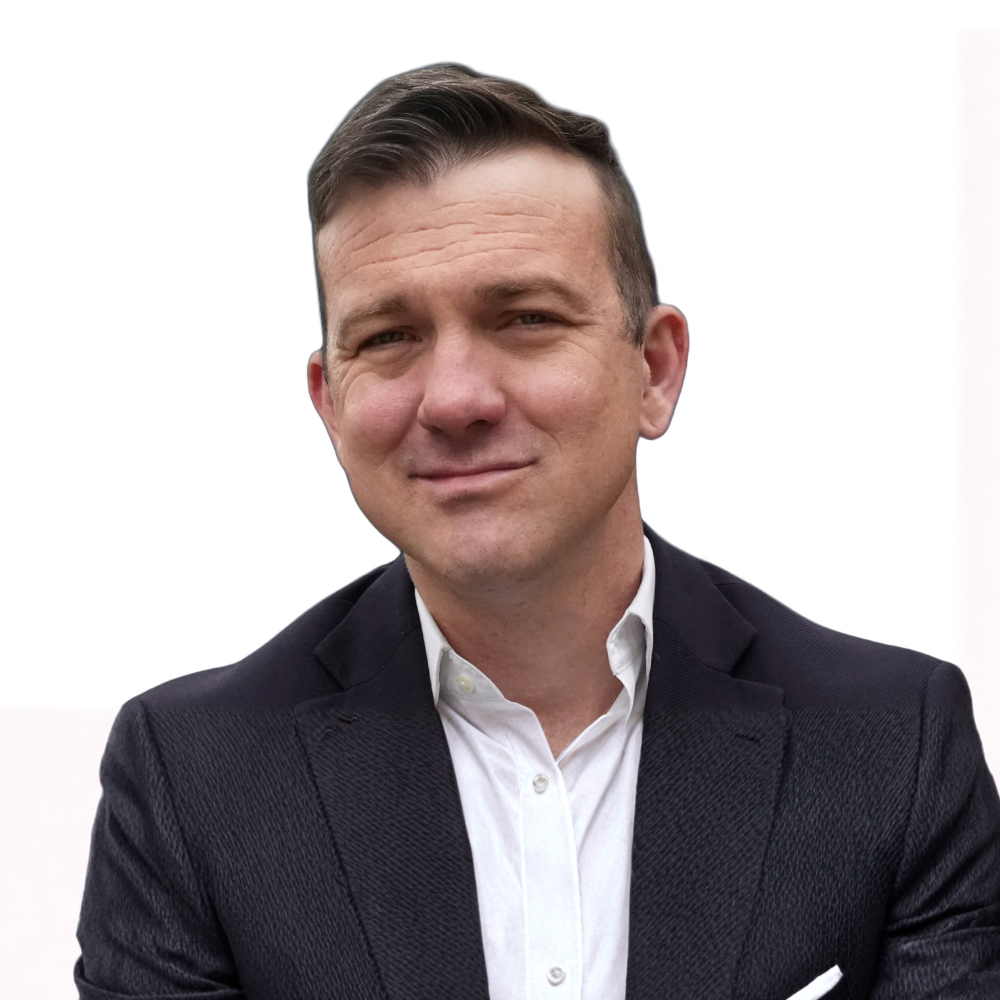 A close-up portrait of a man with short brown hair, wearing a black suit jacket and a white collared shirt, smiling against a plain white background.