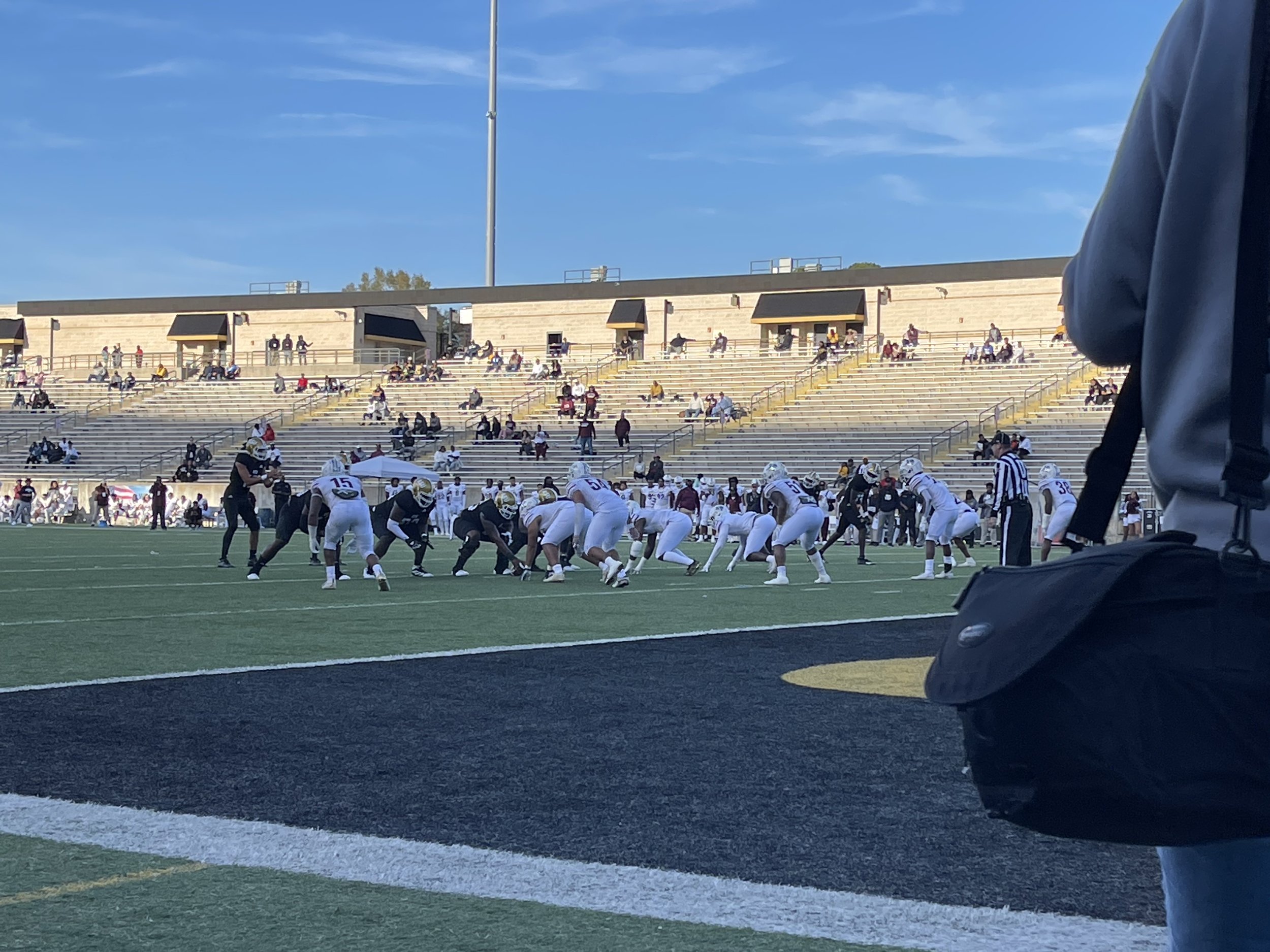 Football game on a field with players in white and black uniforms, referee, and partially filled bleachers.