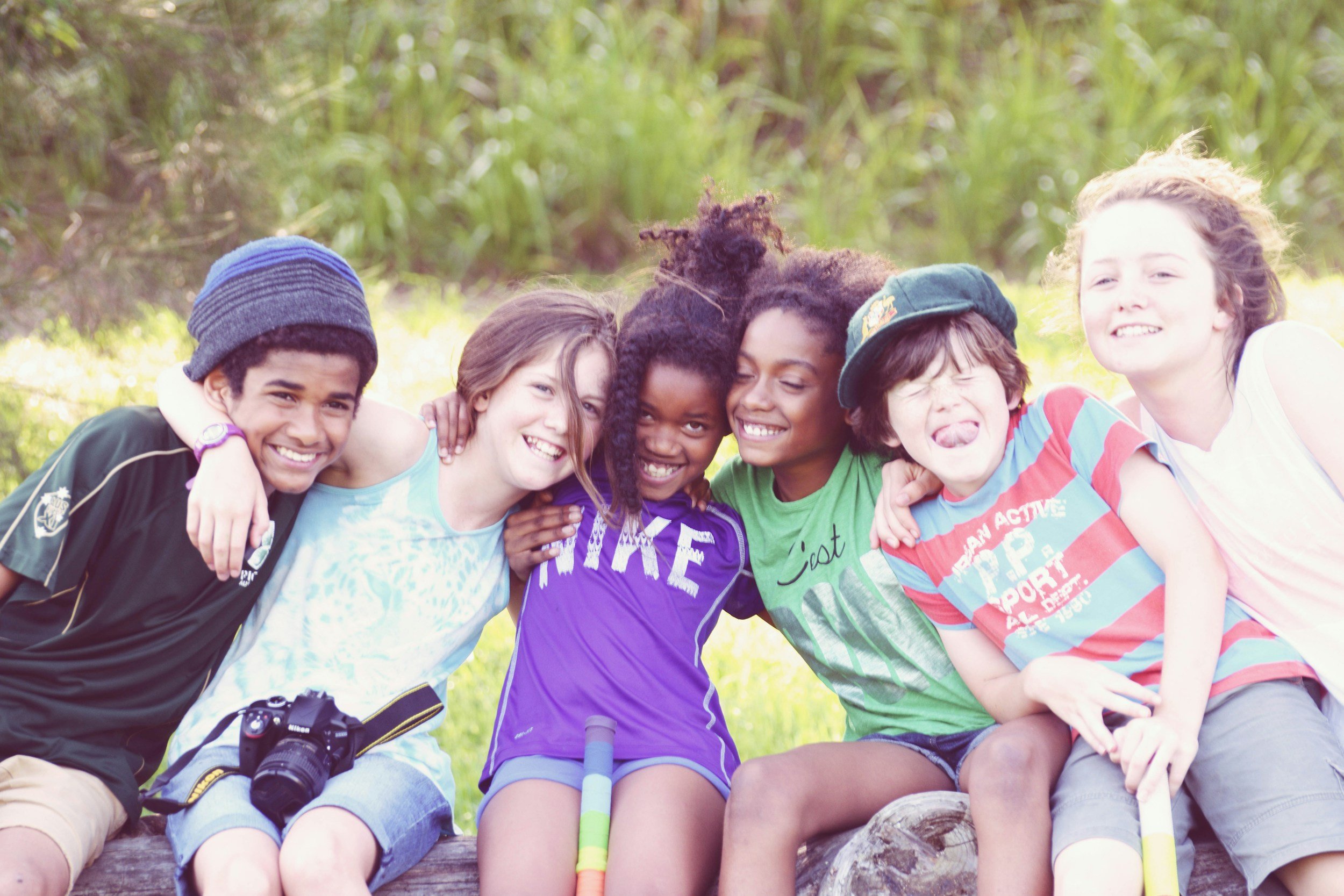 Seven children sitting closely together outdoors with arms around each other, smiling and enjoying a sunny day in a natural setting.