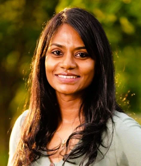 A woman with long dark hair smiling outdoors with a blurred green background.