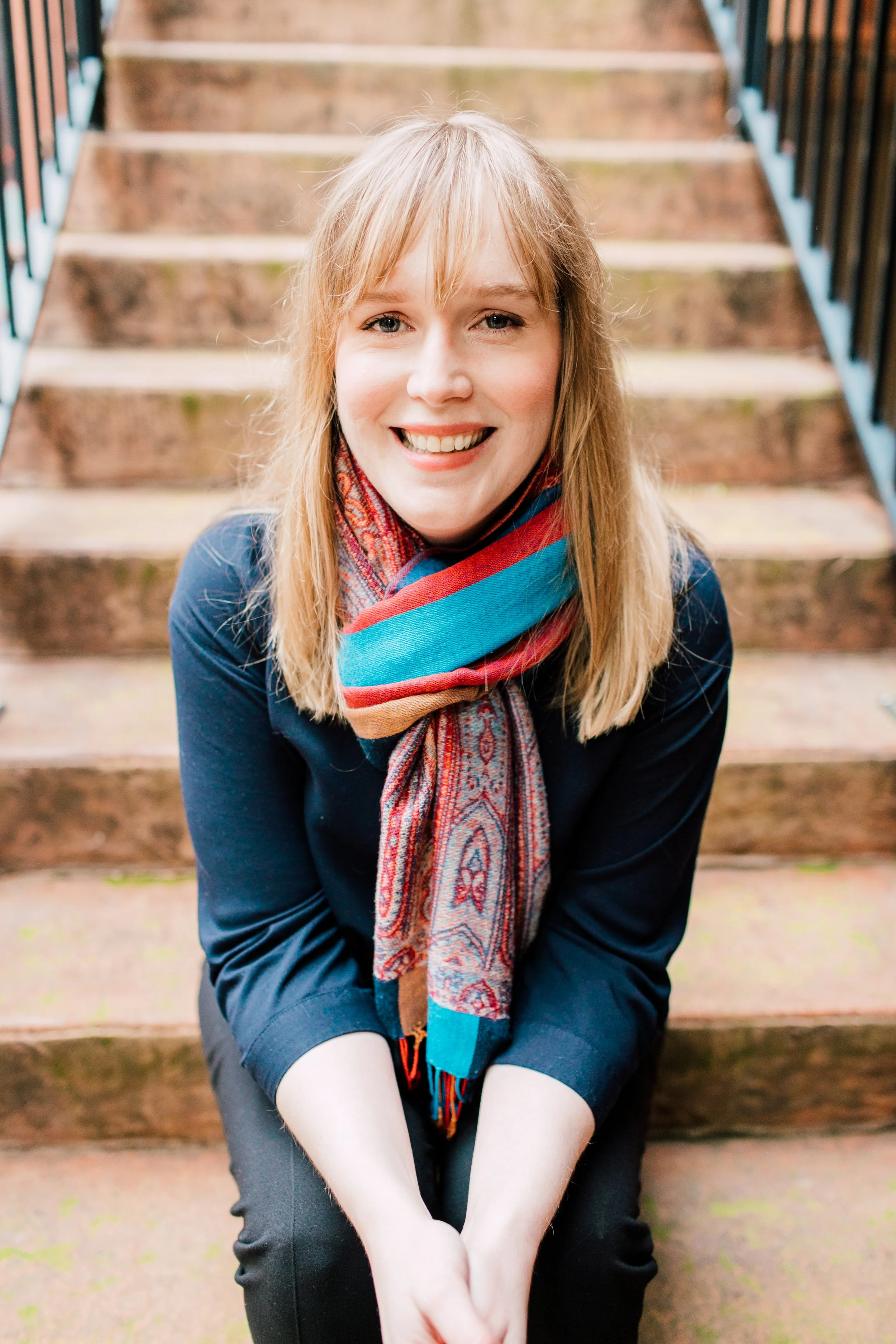 A cheerful blonde woman with blue eyes sitting outdoors on wooden stairs. She is smiling and wearing a colorful scarf and a dark jacket. Her arms are resting on her knees, and she is looking toward the camera.
