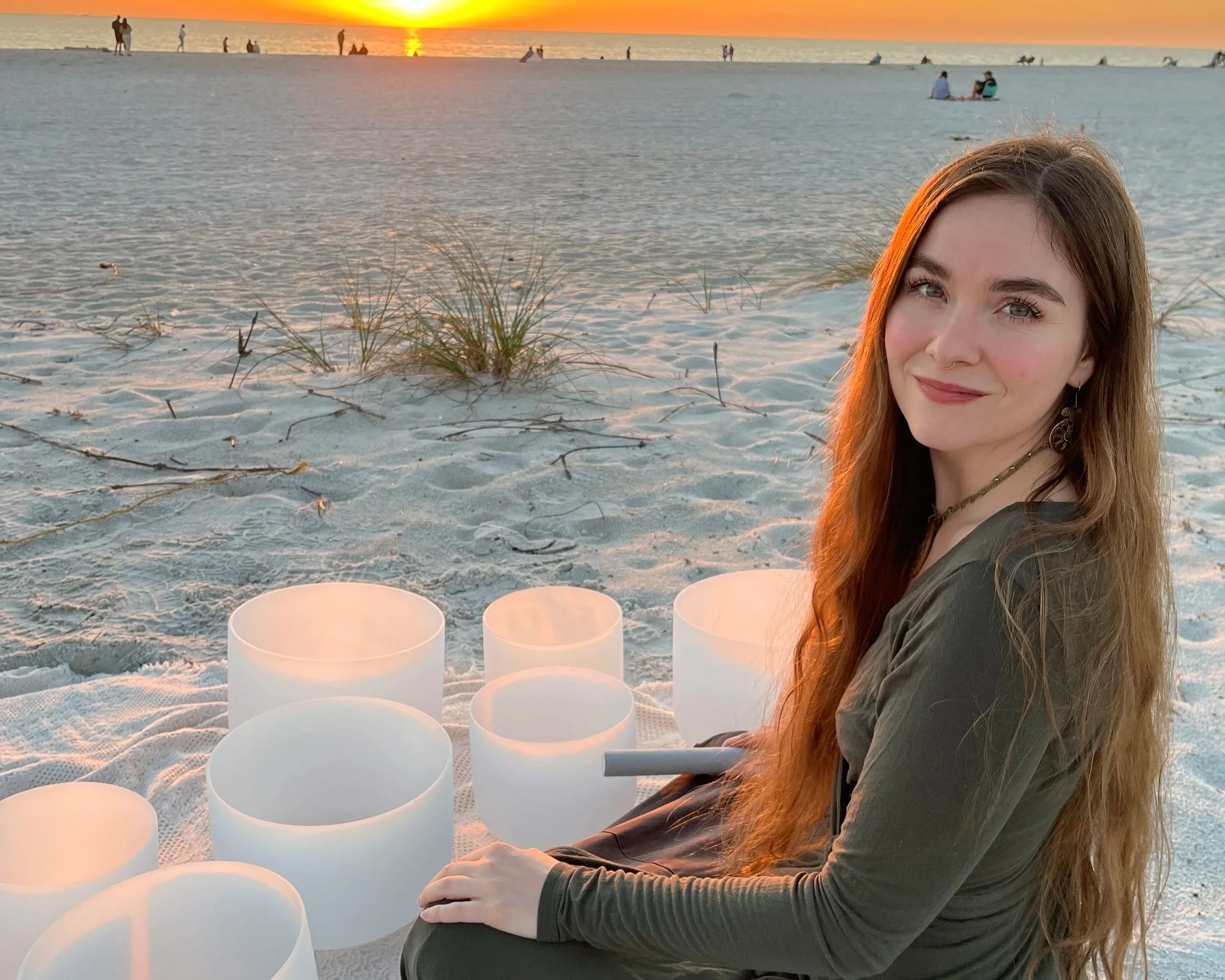 A young woman with long red hair and a necklace sitting on the sand at sunset, with glowing lights in front of her and the ocean in the background.