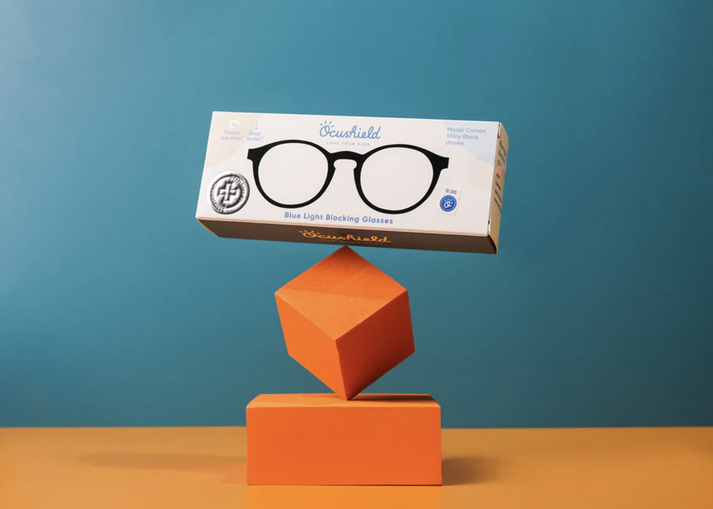 Packaging of blue light blocking glasses balanced on orange geometric shapes against a blue background.