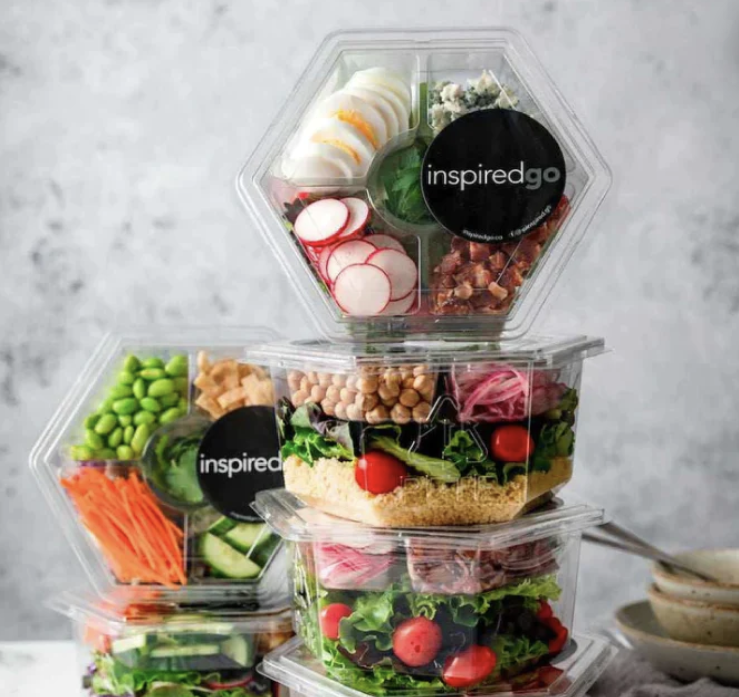 Stacked hexagonal plastic containers filled with various salad ingredients, such as sliced radishes, boiled eggs, edamame, chickpeas, cherry tomatoes, and leafy greens.