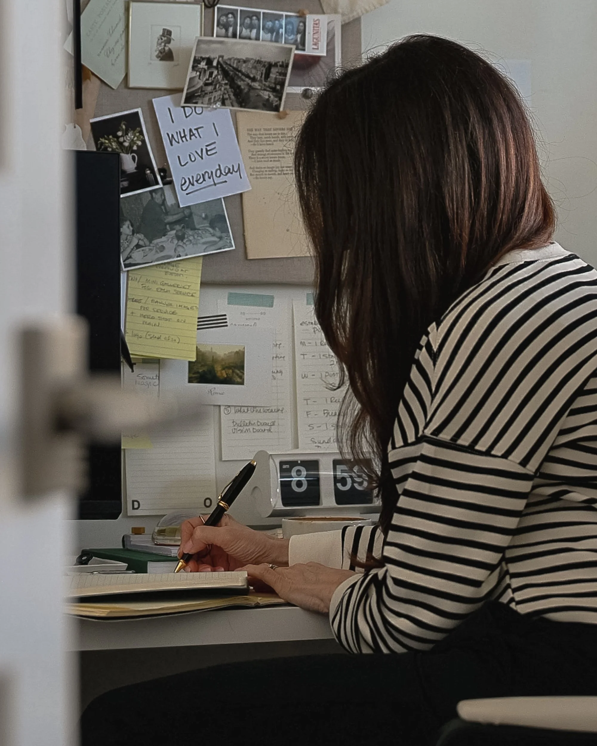 A woman sits at a desk writing in a notebook with a black and gold pen beside a wall filled with photographs, notes, and inspiration clippings. The softly lit workspace and candid composition create an intimate portrait of a quiet creative routine.