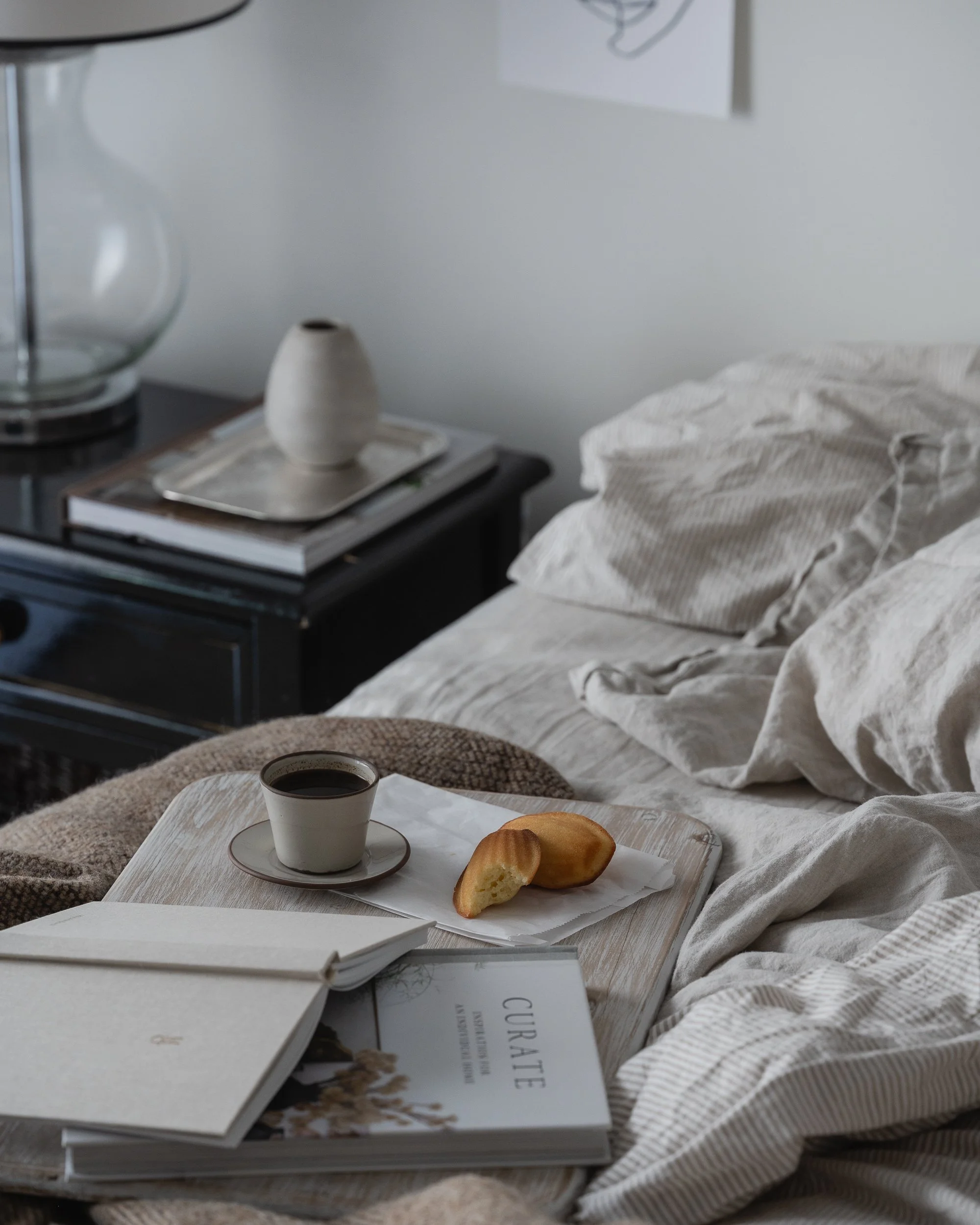 Moody morning bedroom scene with rumpled neutral linen bedding, a wooden breakfast tray holding coffee and madeleines, stacked books and a notebook, soft natural window light, and a dark bedside table with a glass lamp and ceramic vase.