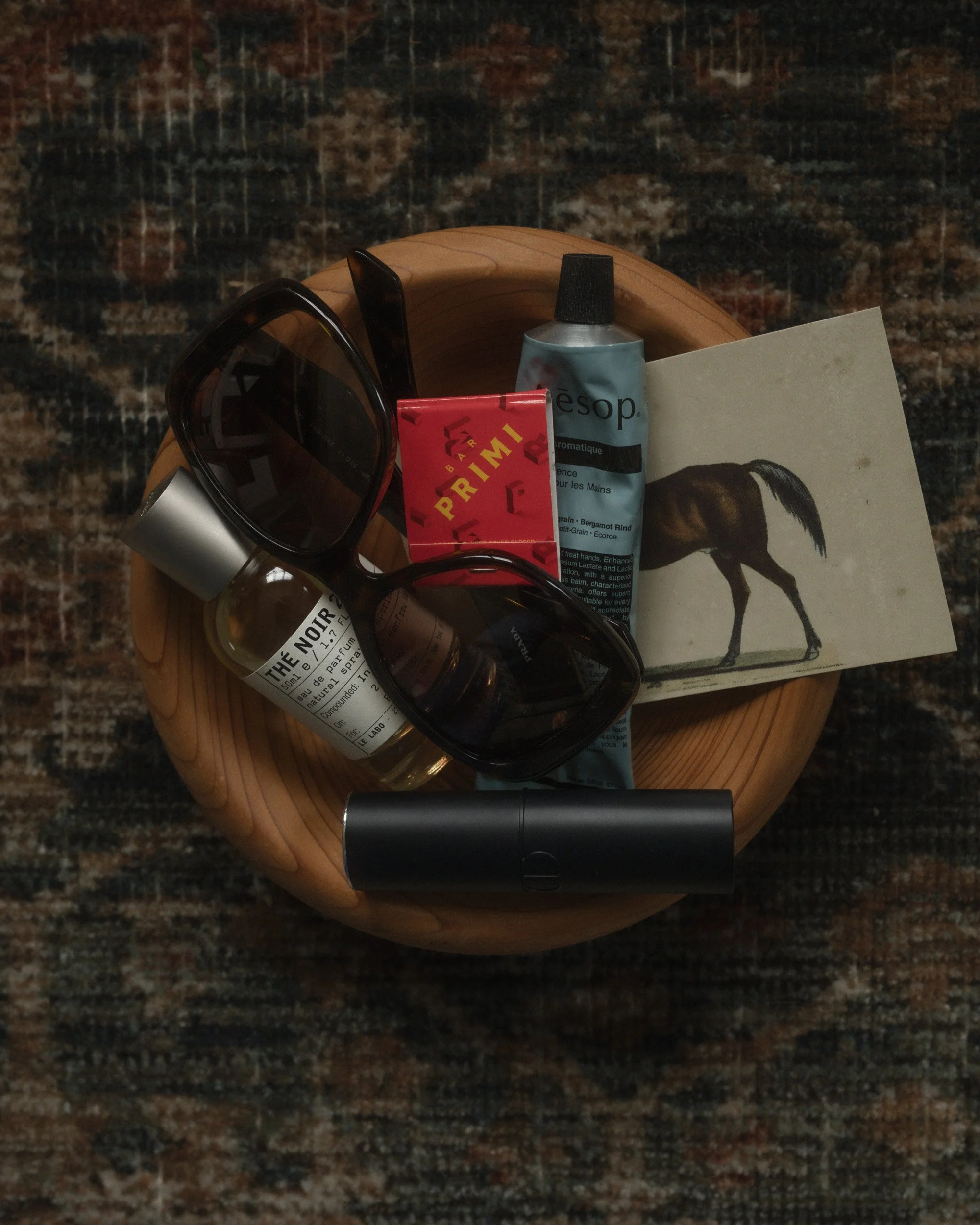 Moody editorial flat lay of everyday objects arranged on a round wooden stool over a patterned rug, including sunglasses, Thé Noir perfume, Aesop hand cream, lipstick, matches, and a horse print card in soft natural light.