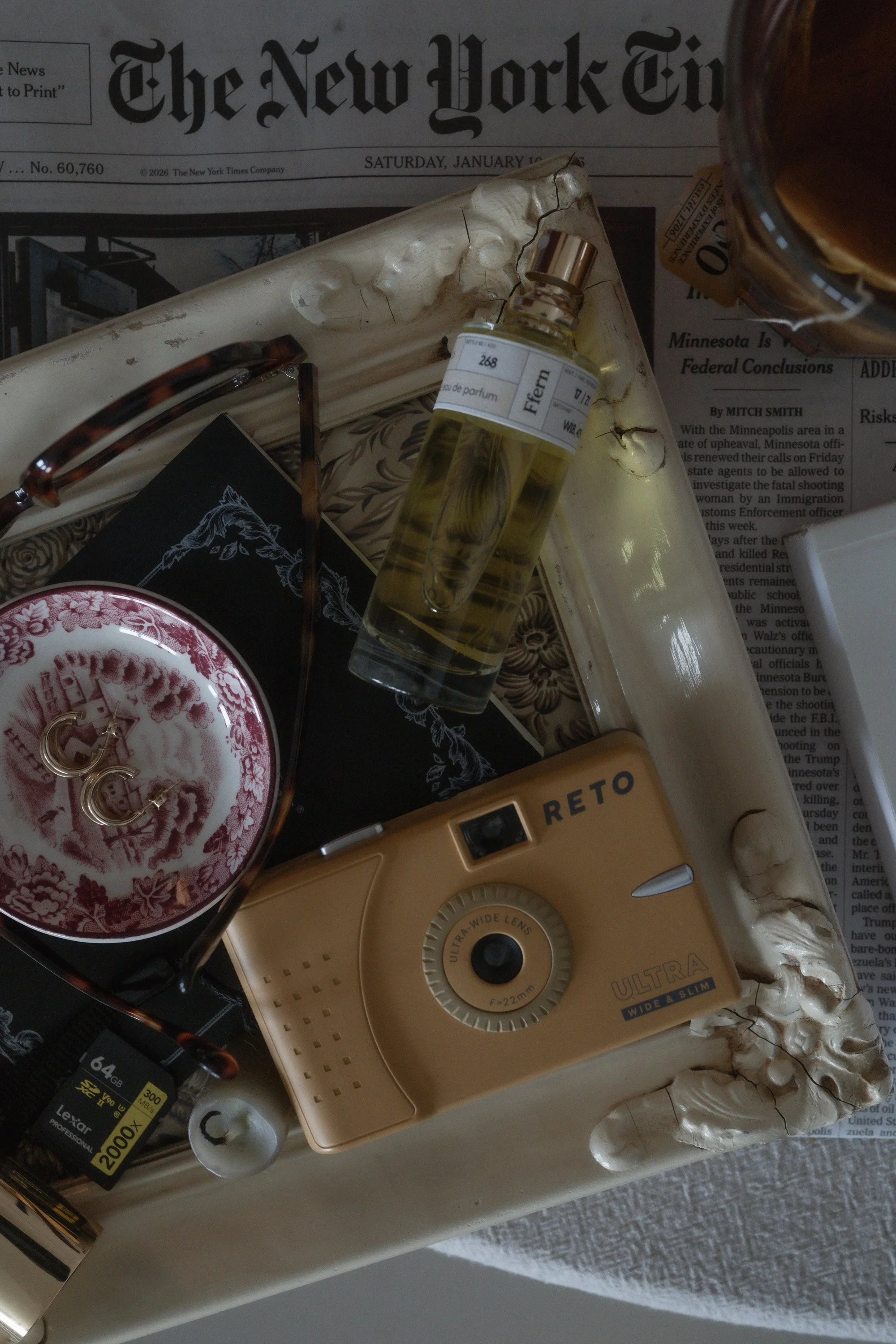 Moody still life flat lay featuring a yellow Reto film camera, Ffern perfume bottle, tortoiseshell glasses, pearl earrings, and small objects arranged inside a vintage ornate frame over a New York Times newspaper.