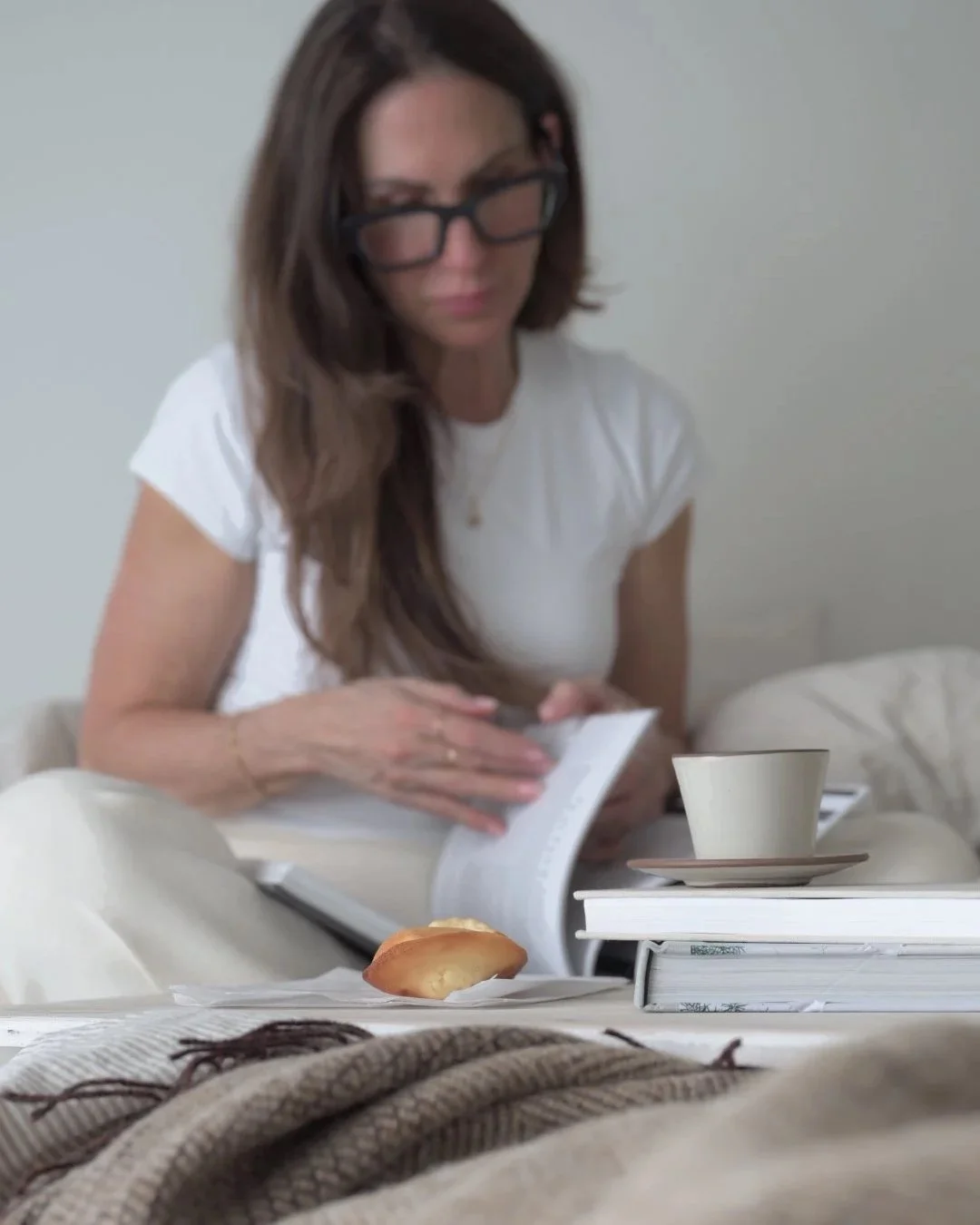 Woman in glasses sitting in an unmade bed reading a book beside a cup of coffee, stacked books, and a pastry, with soft neutral bedding and warm natural light creating a quiet, cozy morning atmosphere.