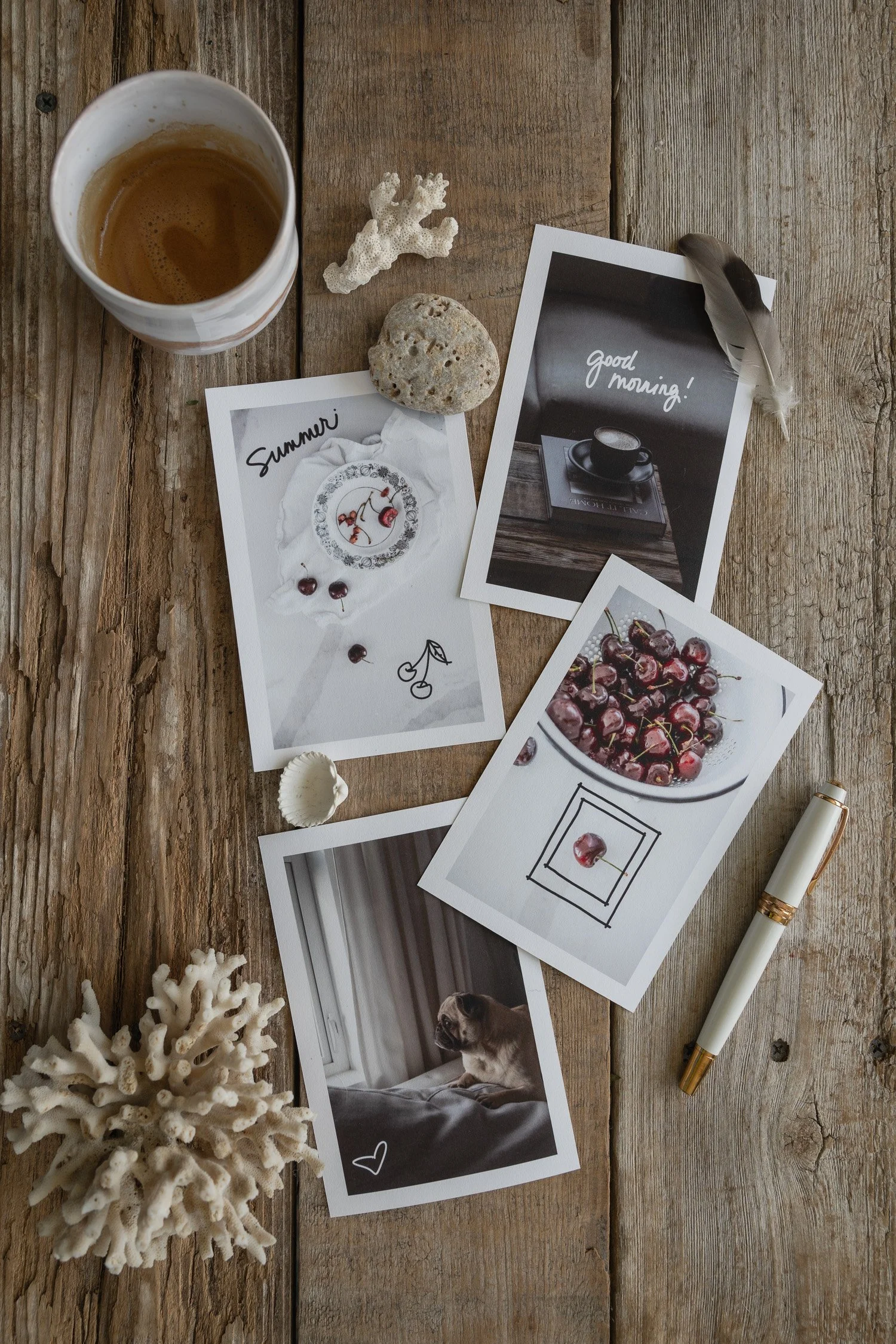 Flat lay of postcard prints featuring lifestyle photographs with handwritten words and simple doodles from the Simple Words & Little Drawings collection, arranged with coffee, a pen, shells, and coral on a rustic wooden table.