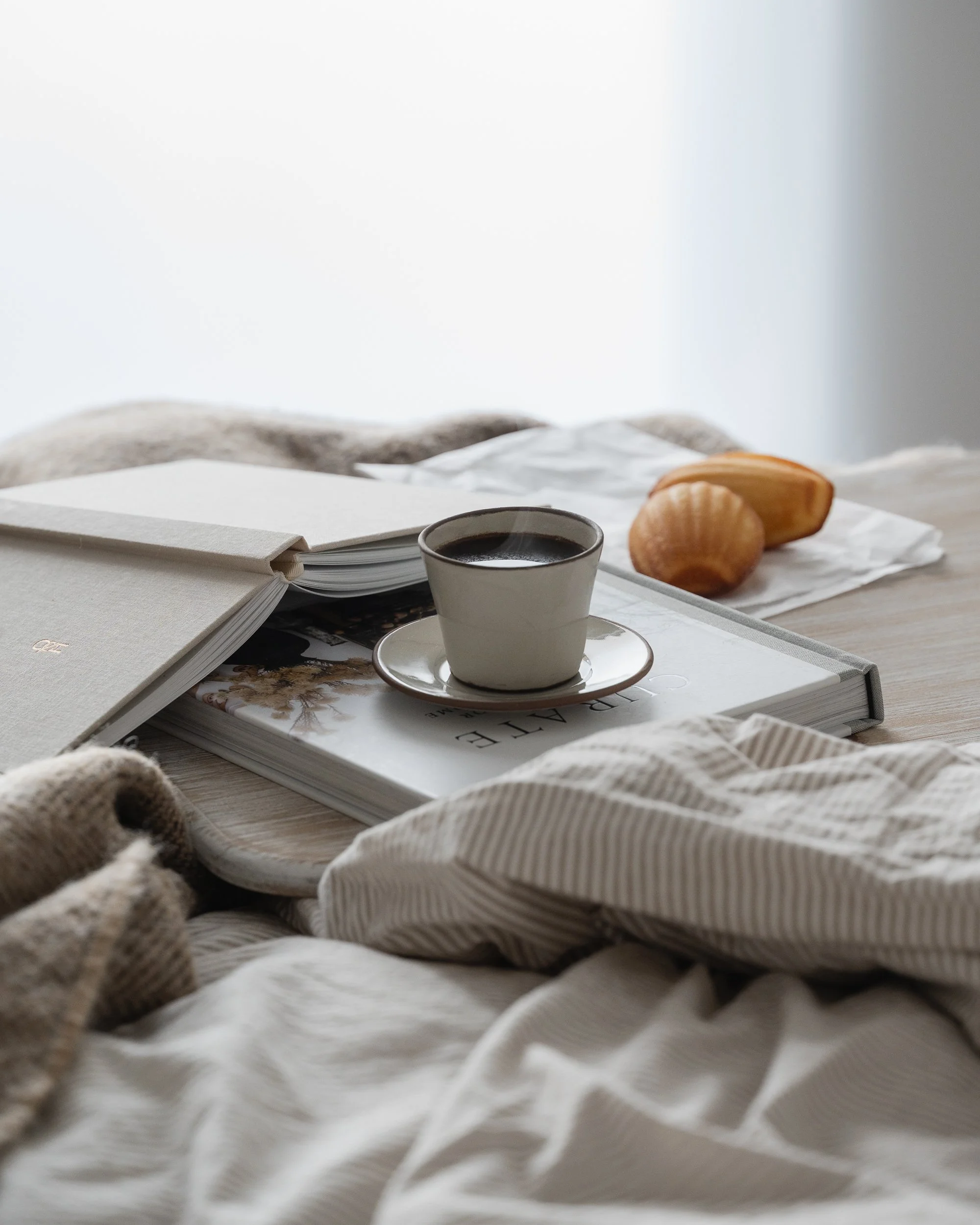 Still life of a ceramic cup of black coffee, madeleine cookies, and stacked books on a wooden tray over rumpled neutral linen bedding, lit by soft natural morning light in a calm minimalist bedroom.