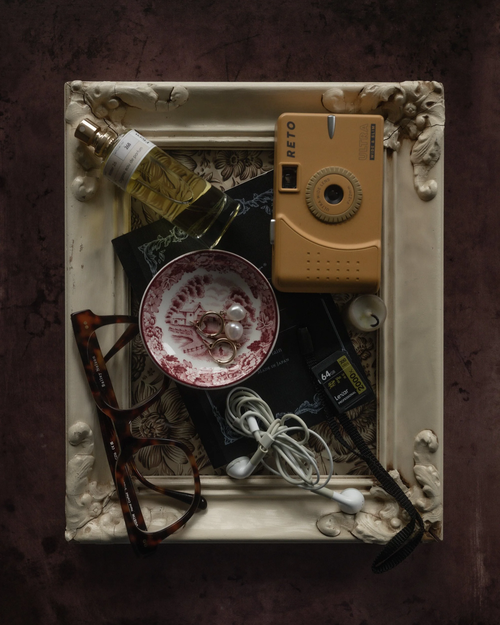 Moody editorial flat lay of personal objects arranged inside a vintage ornate frame, including a yellow Reto film camera, perfume bottle, tortoiseshell glasses, pearl earrings, earbuds, notebook, and memory card on a dark textured background.