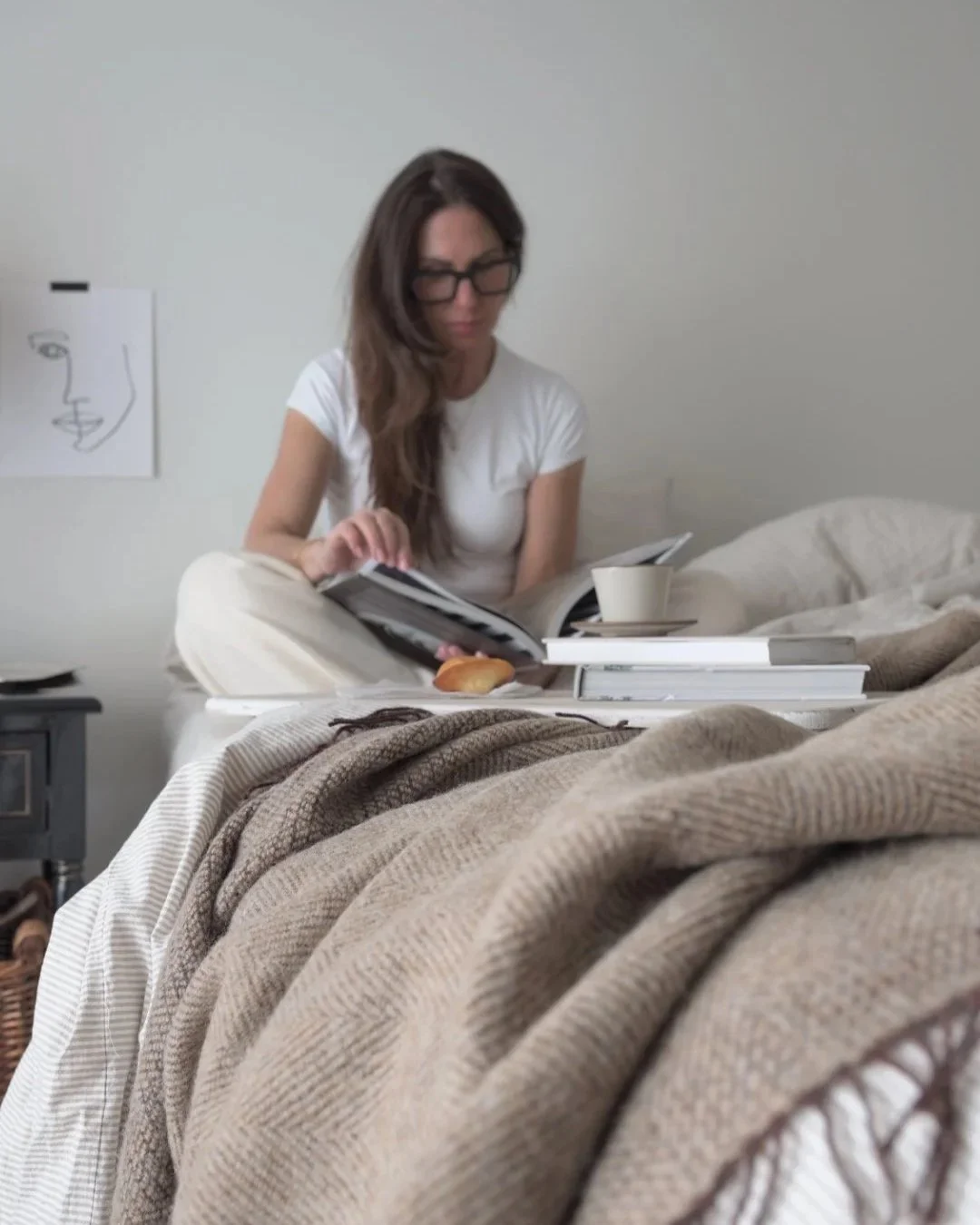 Woman sitting in an unmade bed reading a book beside a cup of coffee and pastry, surrounded by neutral linen bedding and textured blankets in a calm, softly lit minimalist bedroom.