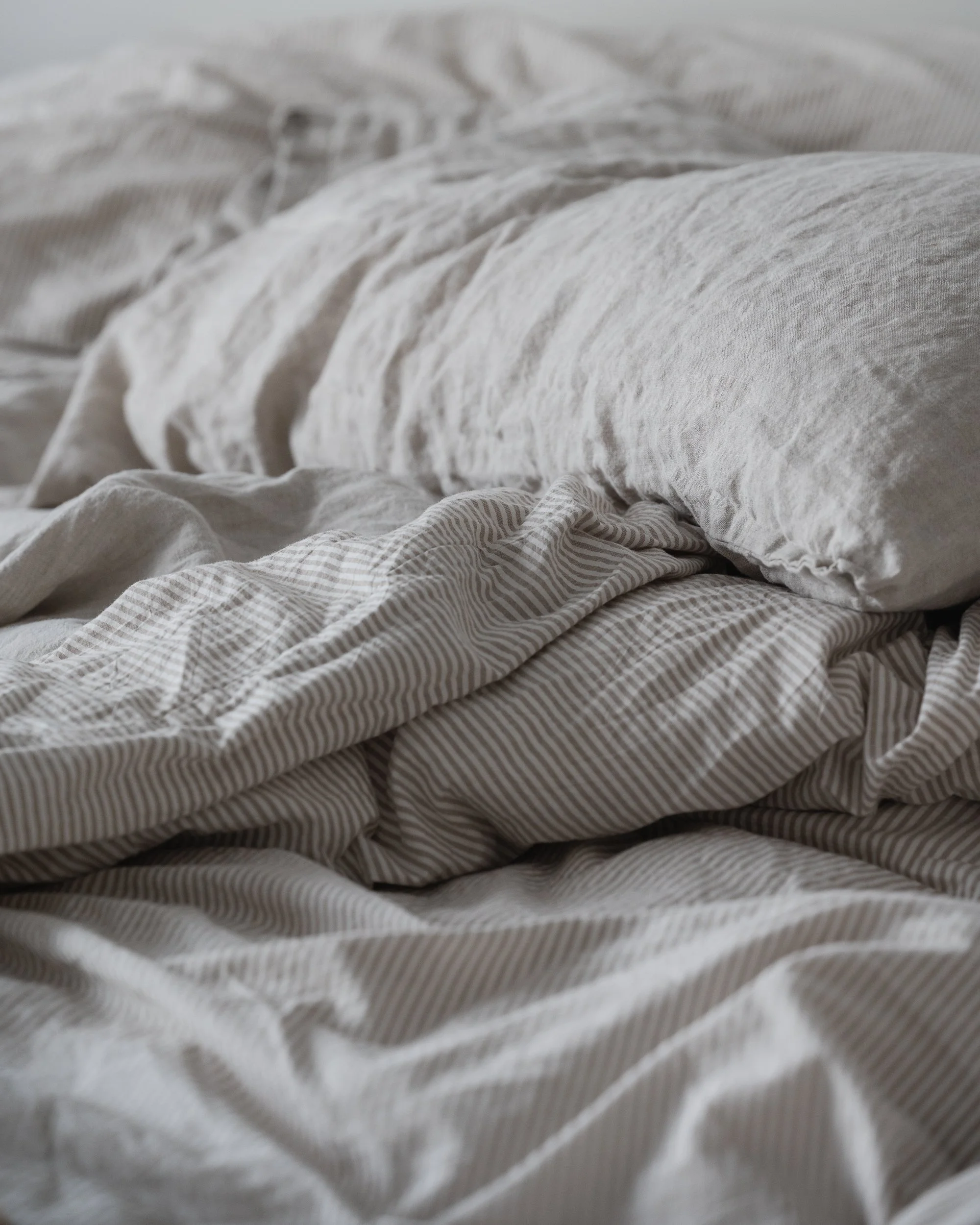 Close-up of rumpled neutral linen bedding and striped sheets in soft natural light, creating a calm, cozy, minimalist bedroom atmosphere with layered texture and muted tones.