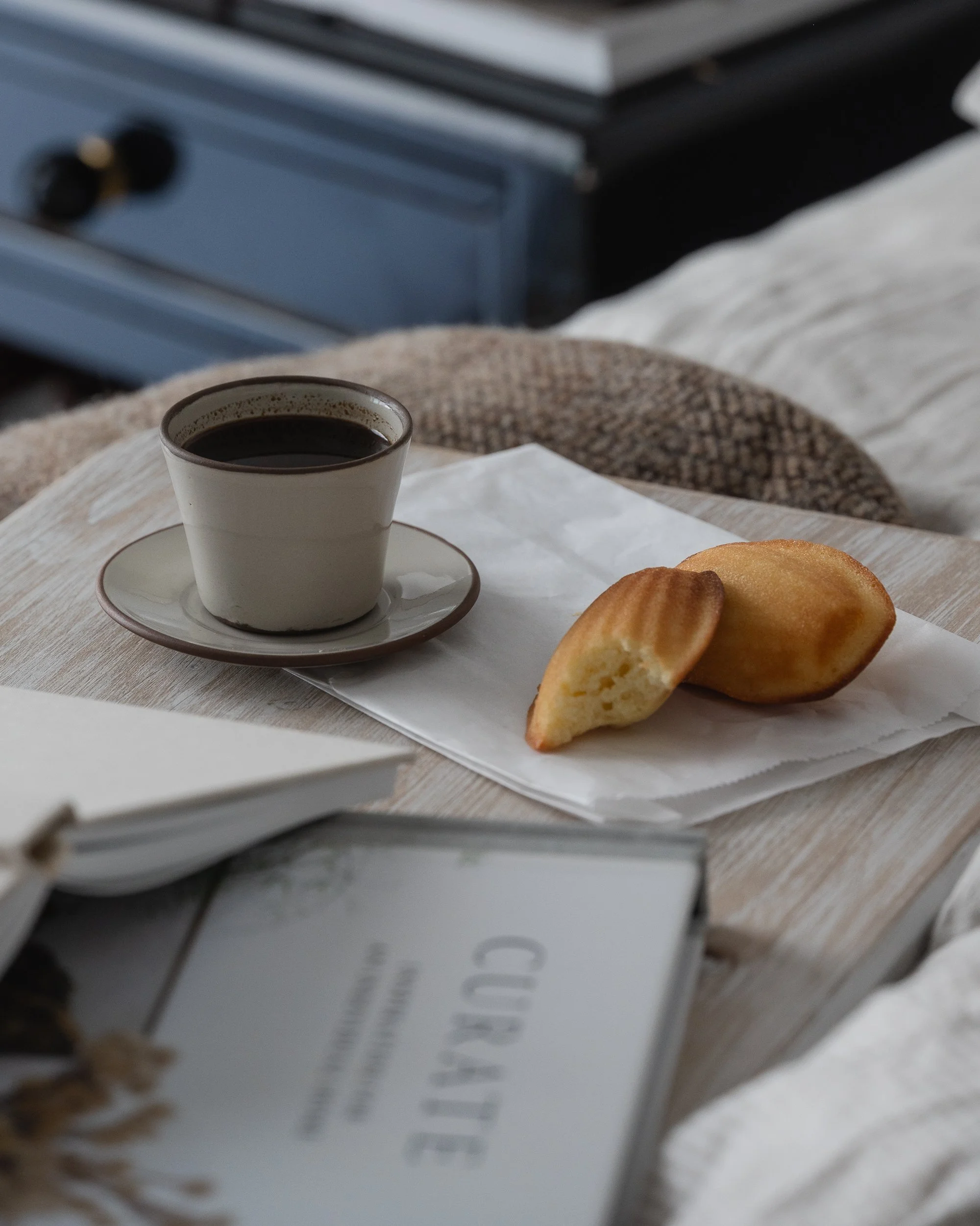 Close-up of a ceramic cup of black coffee and madeleine cookies on a wooden breakfast tray in bed, surrounded by neutral linen bedding, textured blankets, and softly blurred books in warm natural light.
