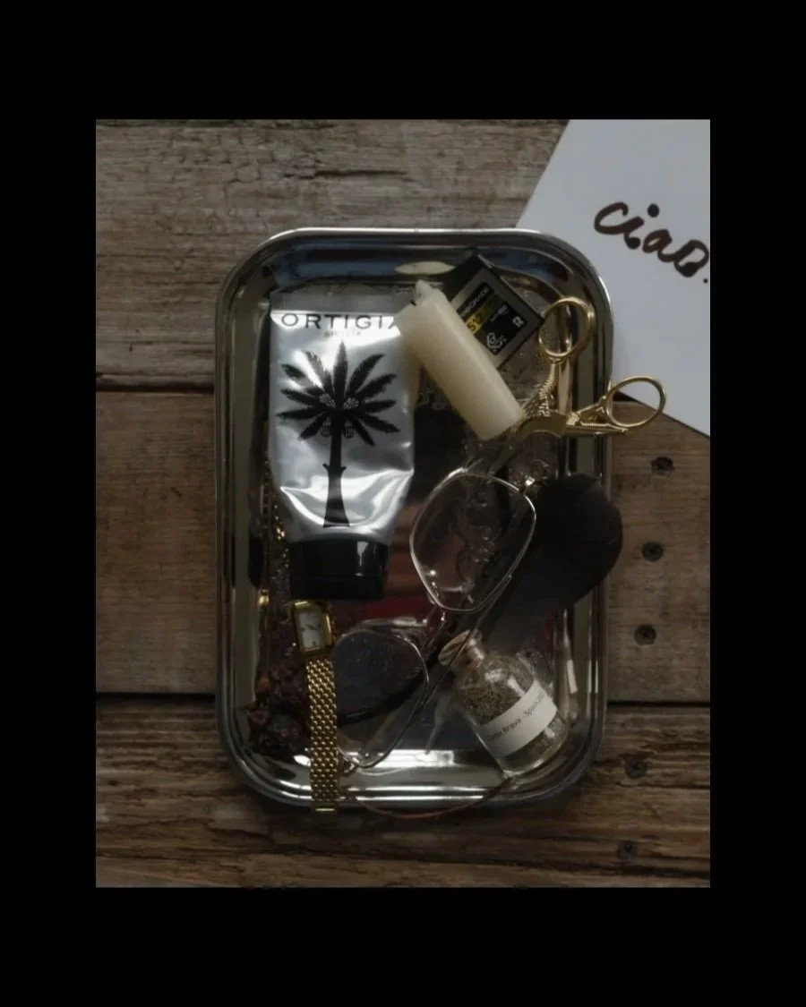 Moody flat lay of personal objects arranged in a silver tray on a rustic wooden surface, including sunglasses, a silver hand cream tube, gold scissors, a watch, candle, film memory card, and small glass bottle beside a handwritten “ciao” card.