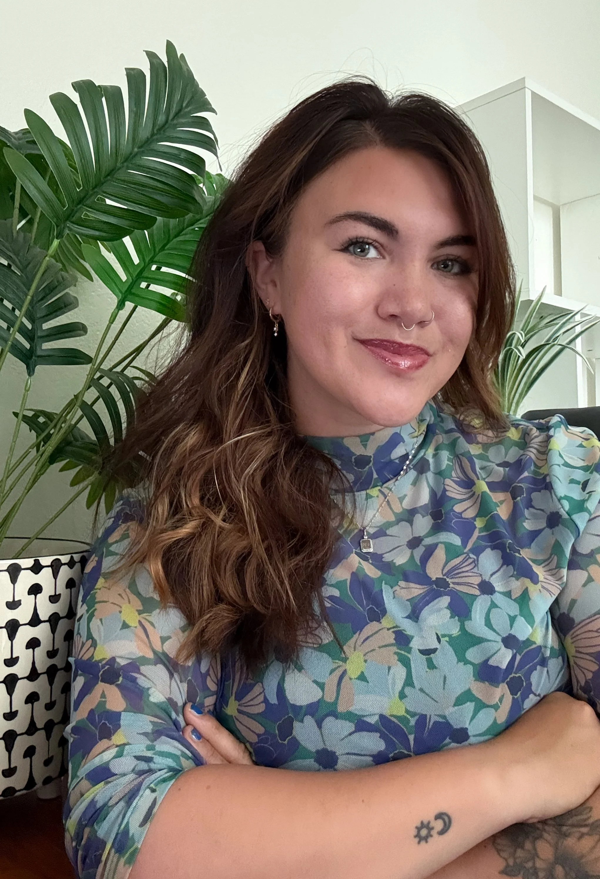 A woman with long wavy hair, light skin, wearing a colorful floral top, with a nose ring, earrings, and a tattoo on her forearm, sitting next to large green indoor plants in a room with light-colored walls.