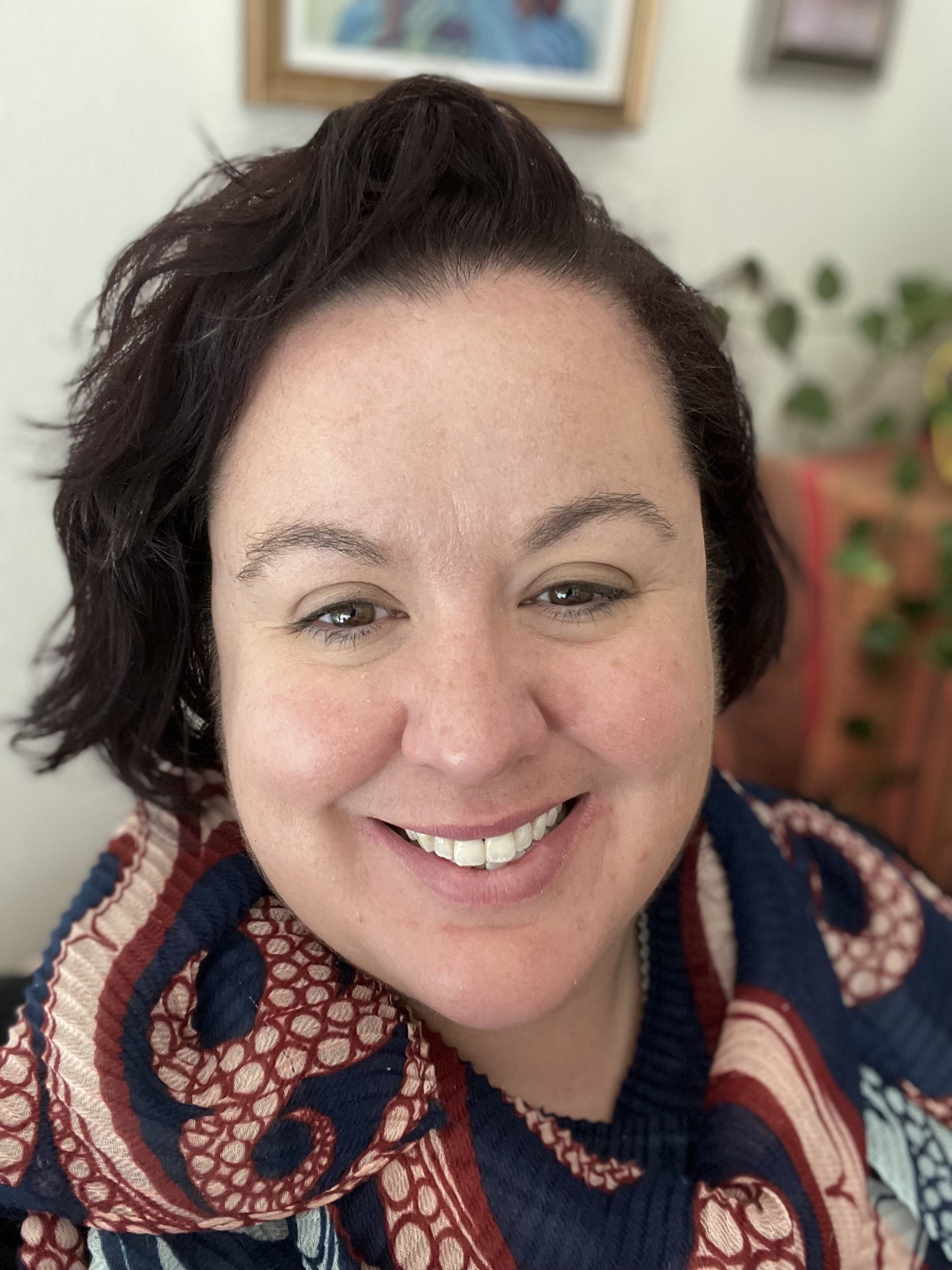 Close-up of a smiling woman with dark wavy hair, wearing a colorful patterned sweater, indoors with artwork and plants in the background.