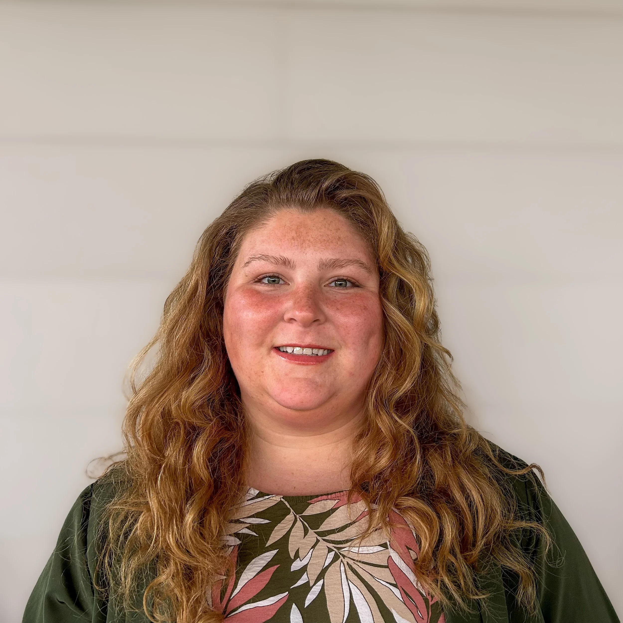 A close-up of a woman's face with reddish-blonde curly hair, smiling, wearing a green jacket and a patterned top with large leaves, standing against a plain background.