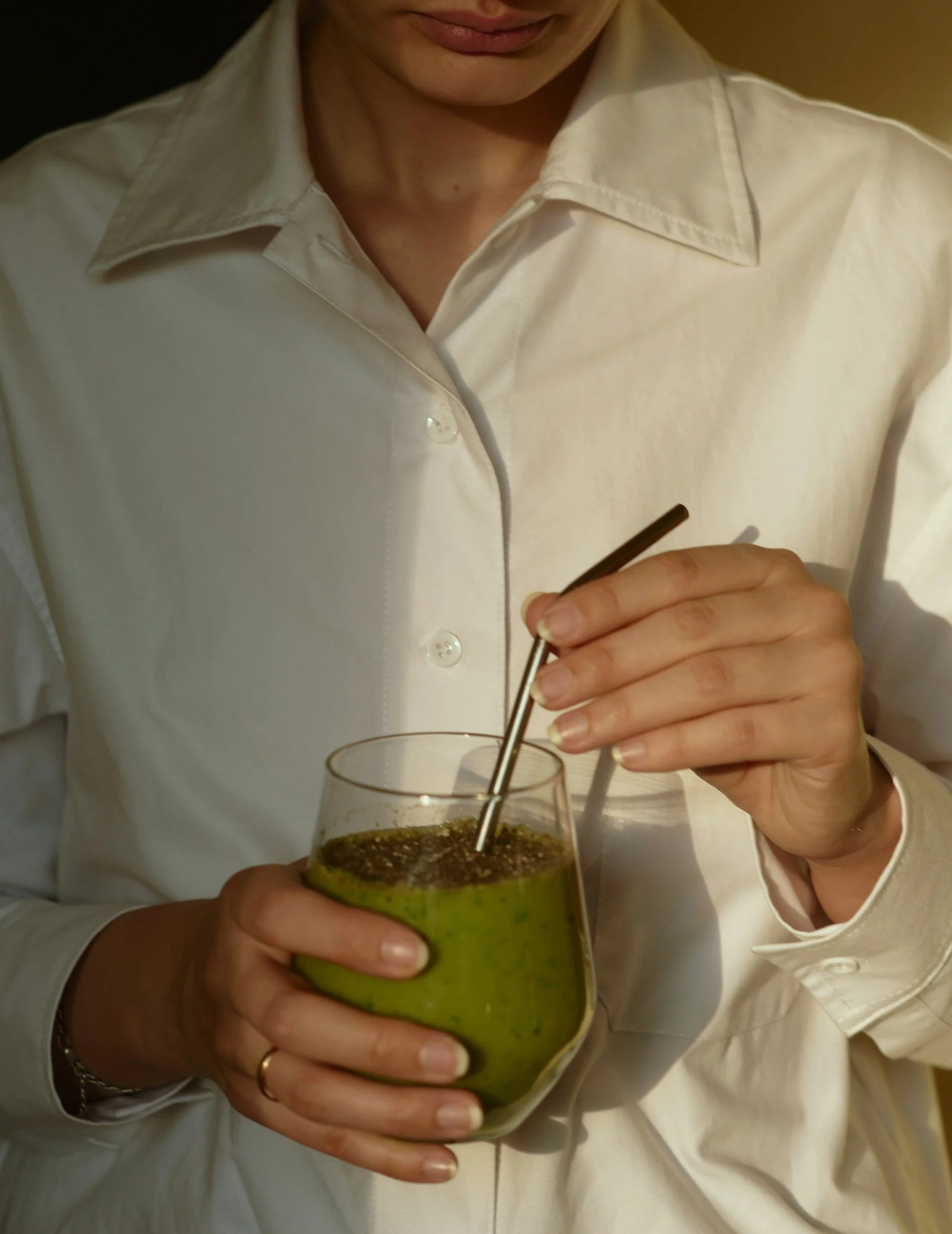 Person wearing a white shirt holding a glass of green smoothie and stirring it with a metal straw.