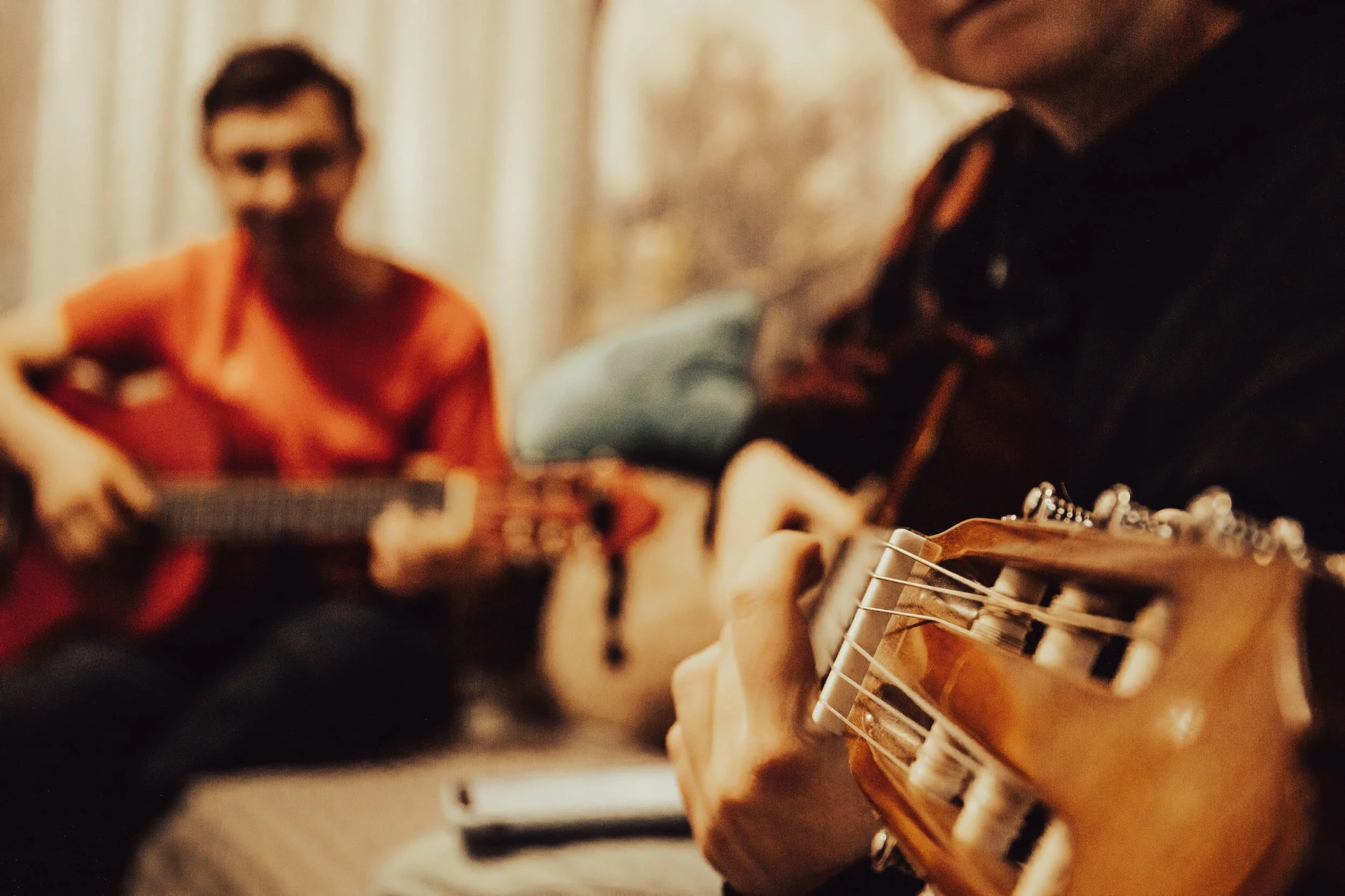 Close-up of a person playing an acoustic guitar with another person in the background holding a guitar in a cozy indoor setting.