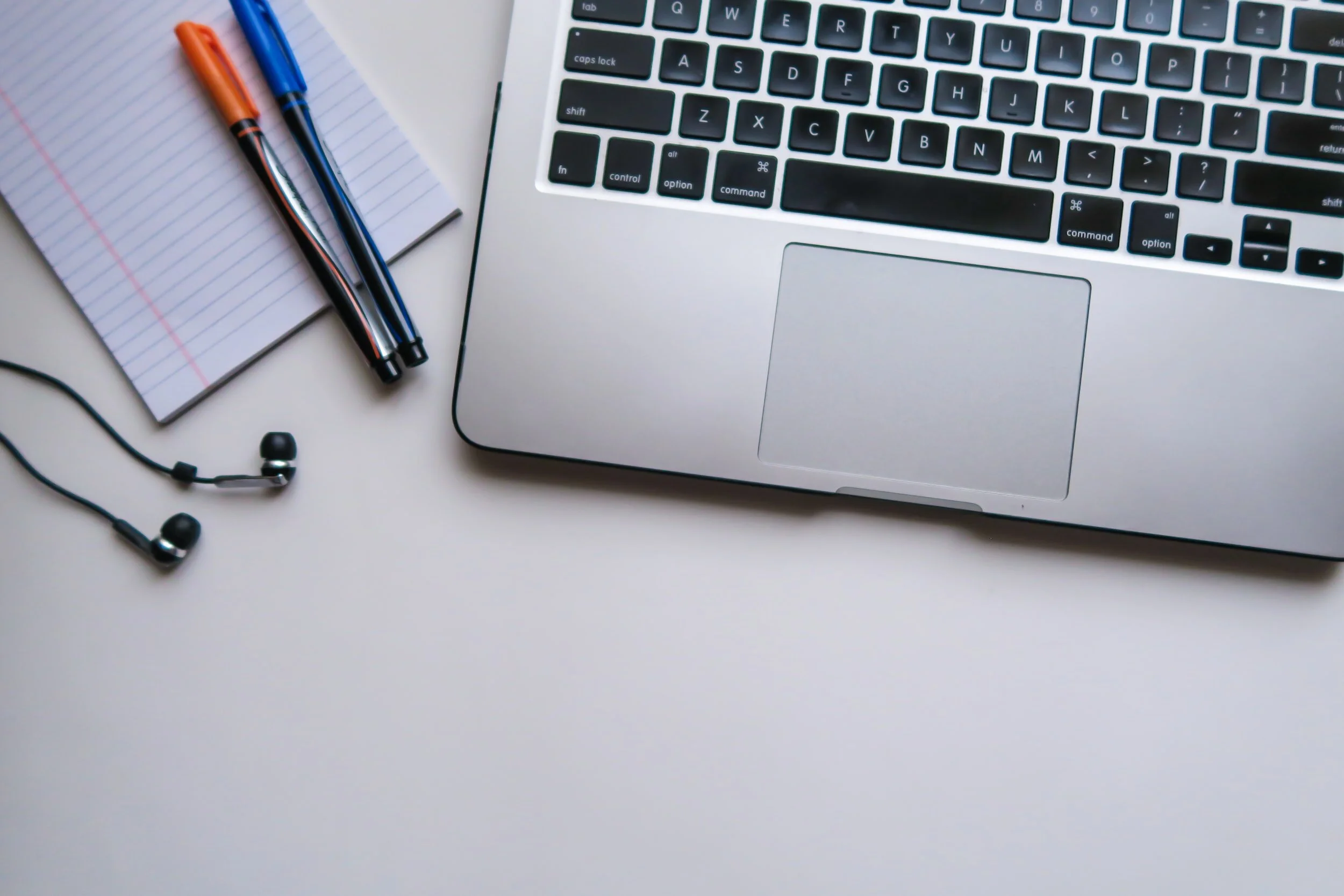 A silver laptop, a small lined notepad, two pens (orange and blue), and black earphones on a white surface.