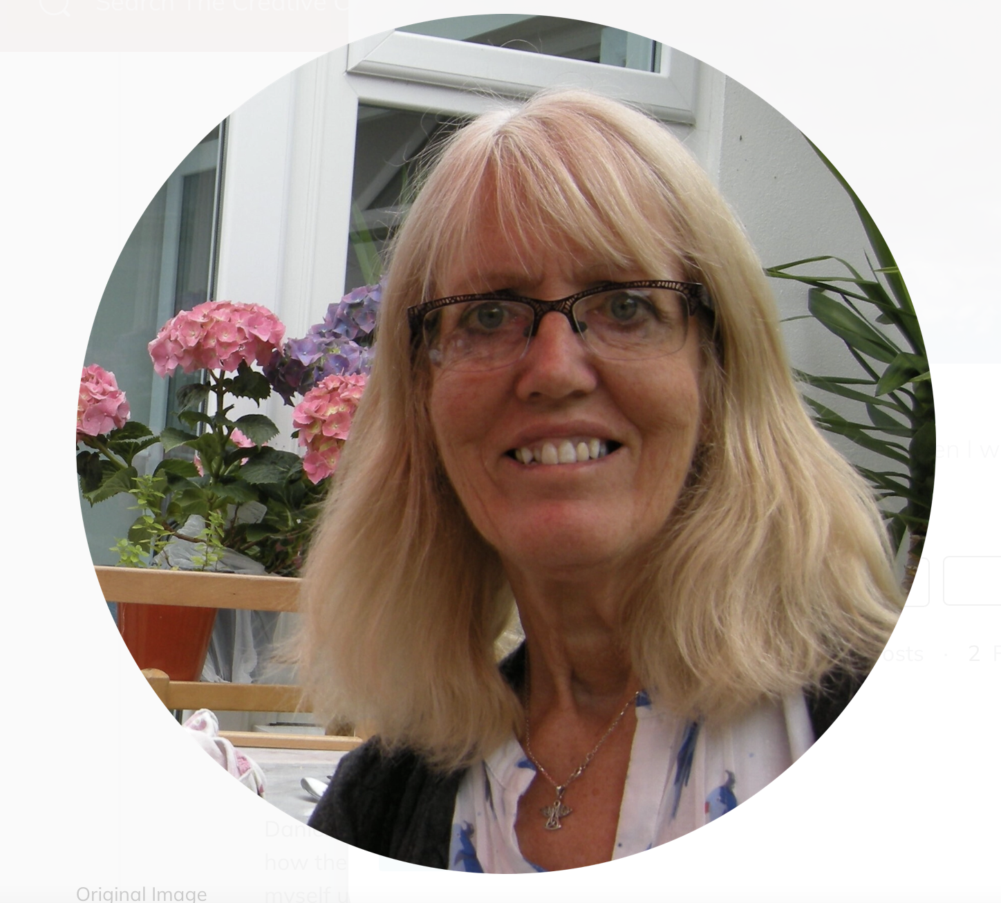 A smiling blonde woman wearing glasses, a necklace, and a white blouse with a black cardigan, in a conservatory or greenhouse with pink and purple hydrangea flowers behind her.