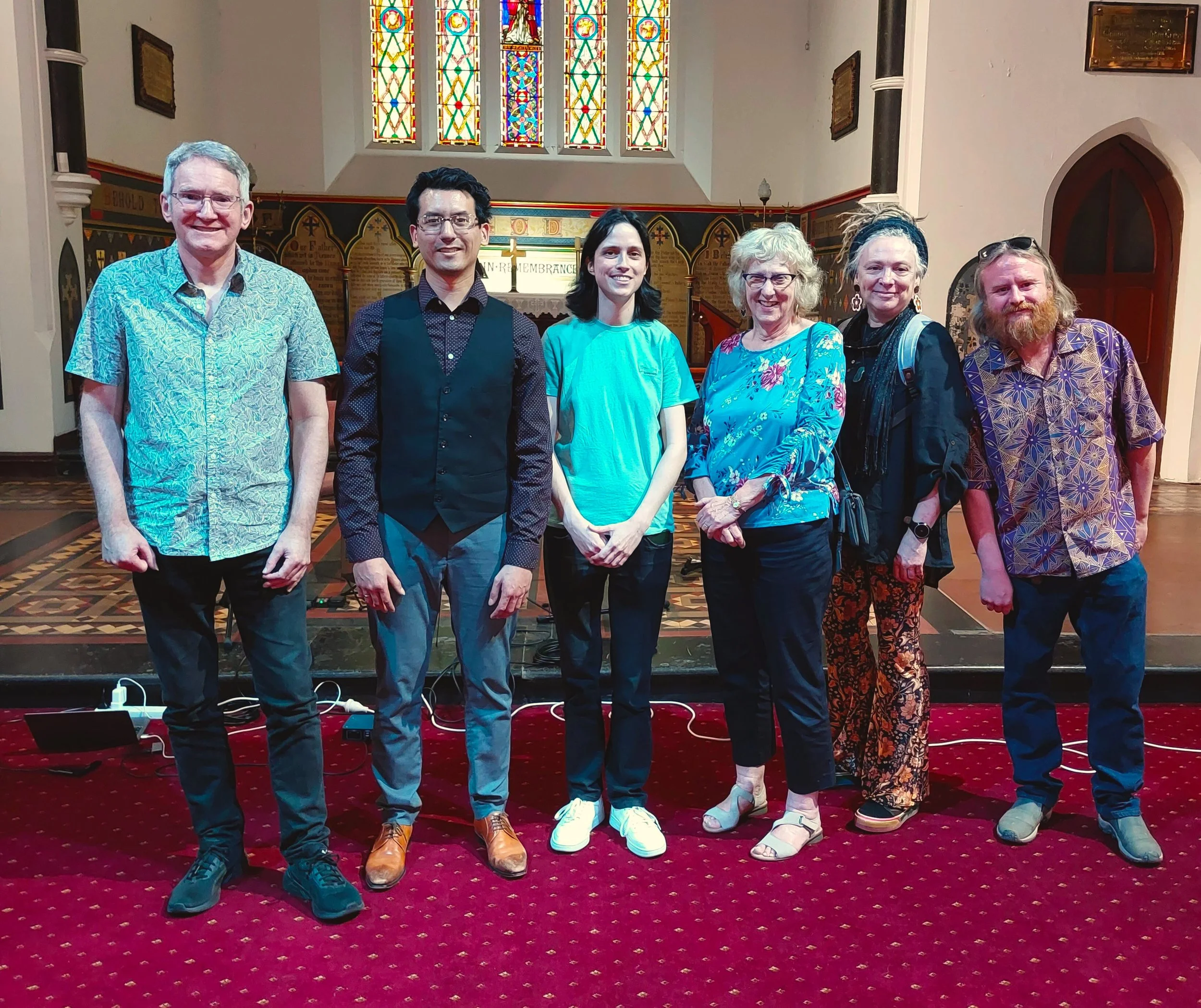 Group of seven people standing inside a church with stained glass windows behind them. They are smiling and dressed in casual to semi-formal attire, standing on a red carpet.