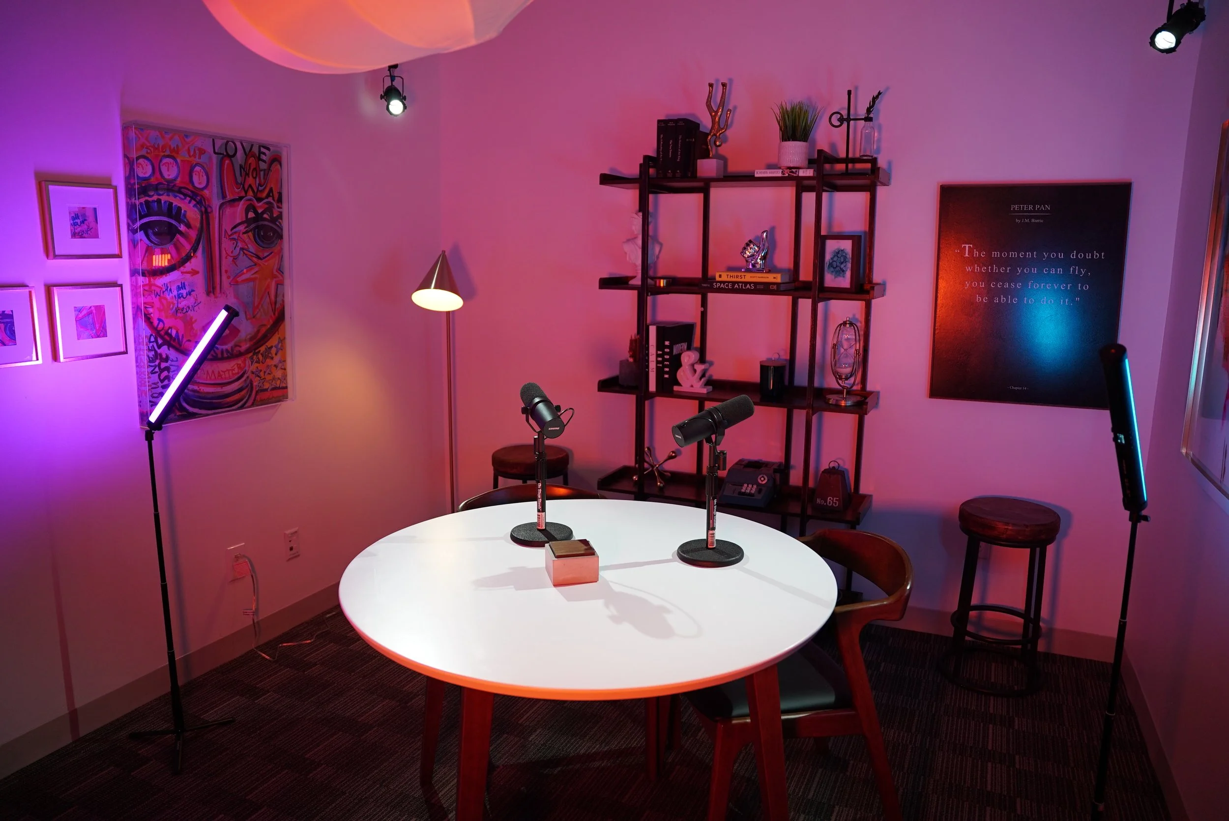 A modern podcast recording room with a round white table, three microphones, and surround lights, decorated with artwork, books, and plants.
