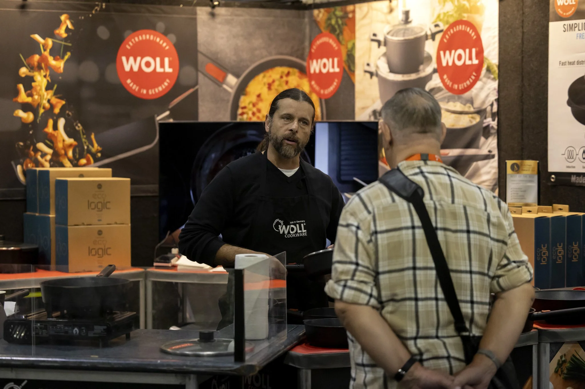 A man with a beard and long hair, wearing a black apron with the WOLL logo, is standing behind a counter talking to another man with glasses and a plaid shirt. Behind them are posters of kitchenware and food items, with the WOLL logo visible in the background.