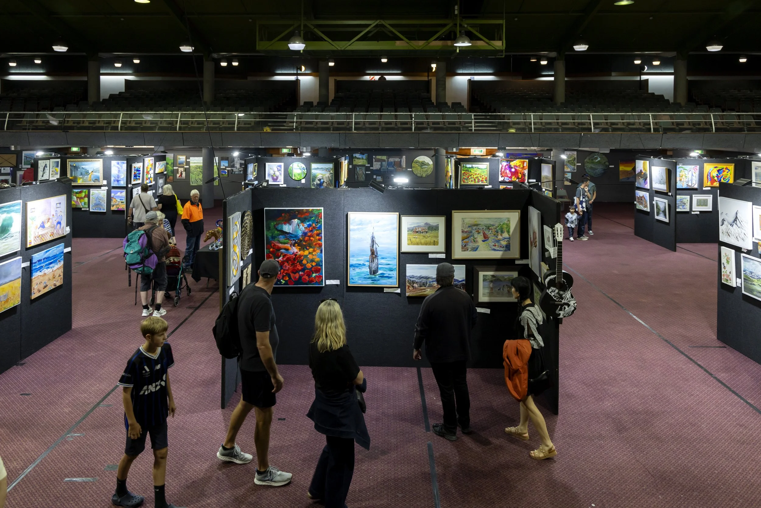 People viewing and walking around an art gallery with colorful paintings displayed on black partitions inside a large indoor space.