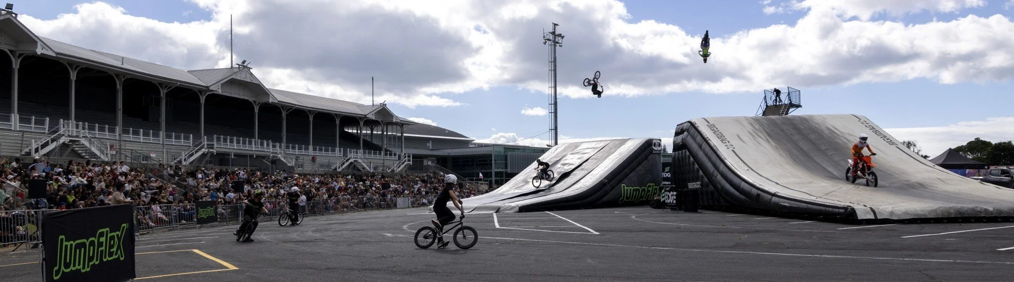 BMX bike riders performing tricks on a large inflatable ramp at a skatepark with a crowd in the stands and people in the background.