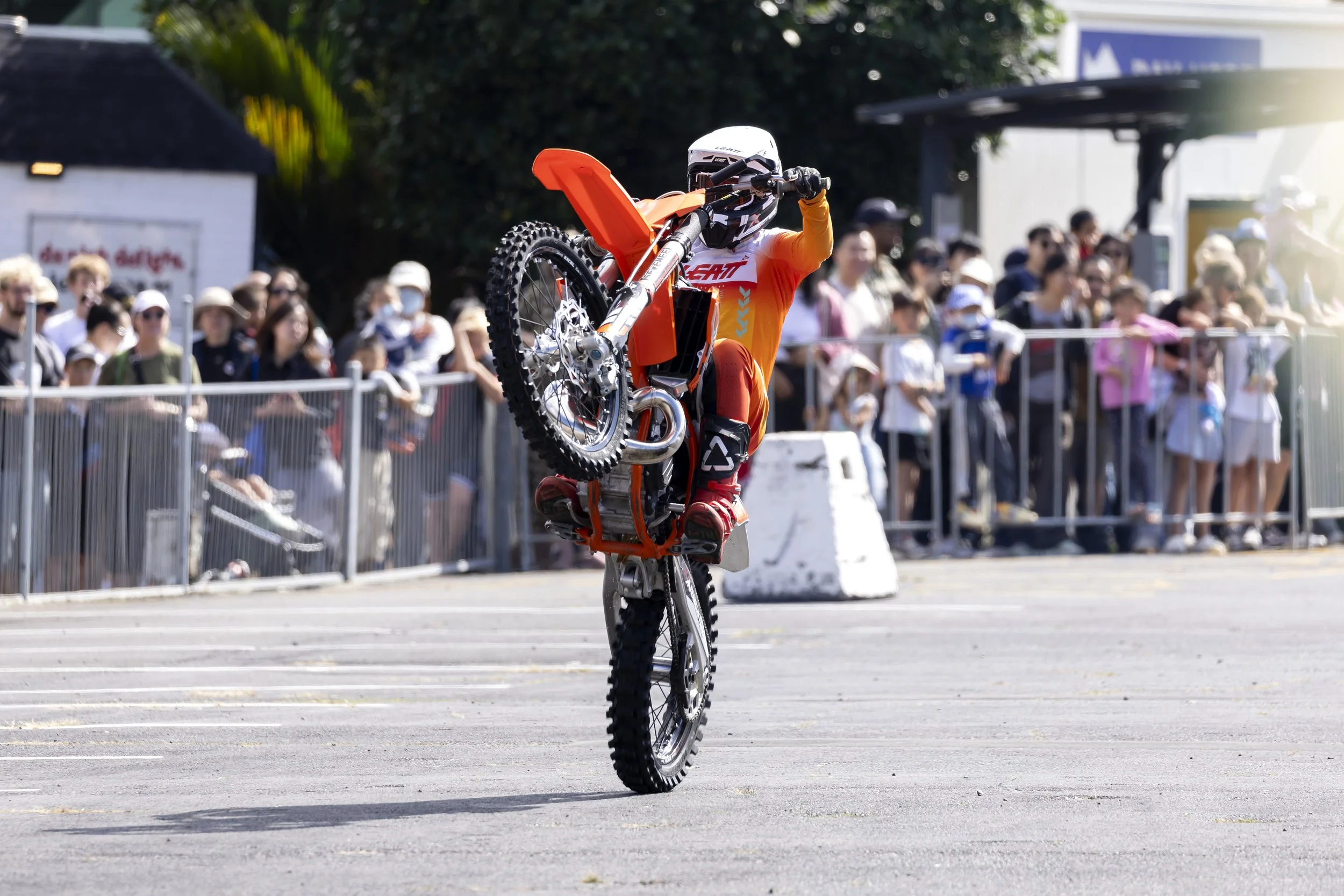 A person performing a wheelie on a dirt bike at an outdoor event, with a crowd of spectators watching behind a metal barricade.