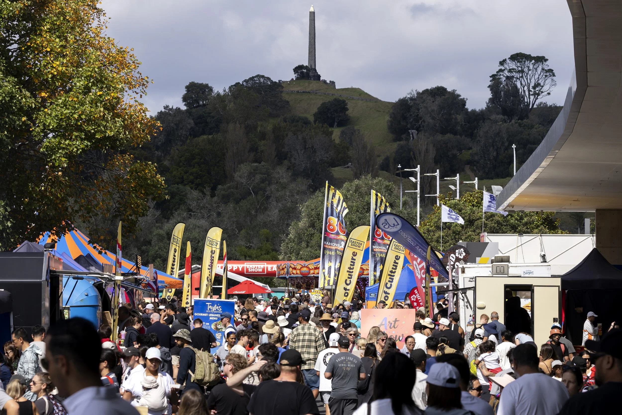 Crowd of people gathered at an outdoor event with colorful banners and tents, with a hill and a tall monument in the background.