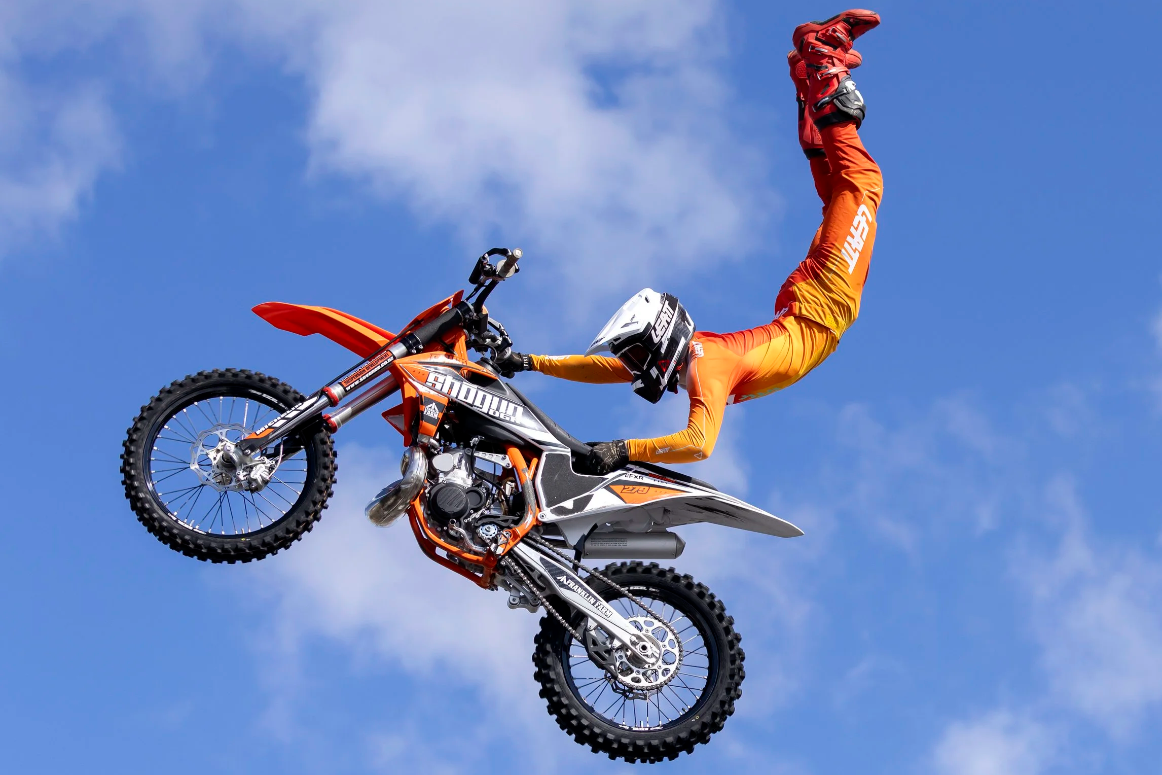 Motocross rider performing a stunt on a dirt bike in mid-air against a blue sky with some clouds.