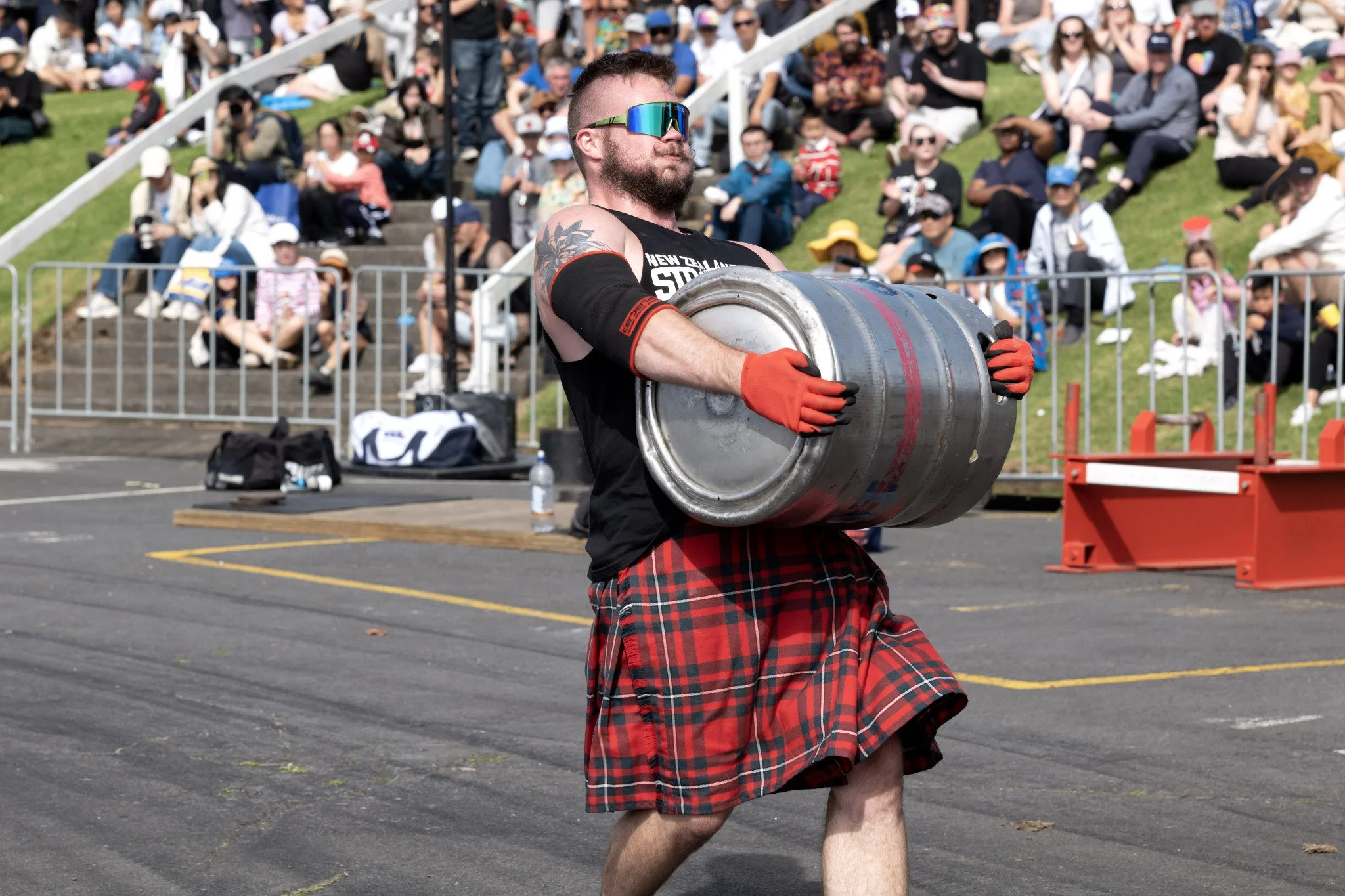 A man wearing sunglasses, gloves, a sleeveless shirt, and a tartan kilt is holding a large metal keg at an outdoor event with a crowd sitting on grass in the background.