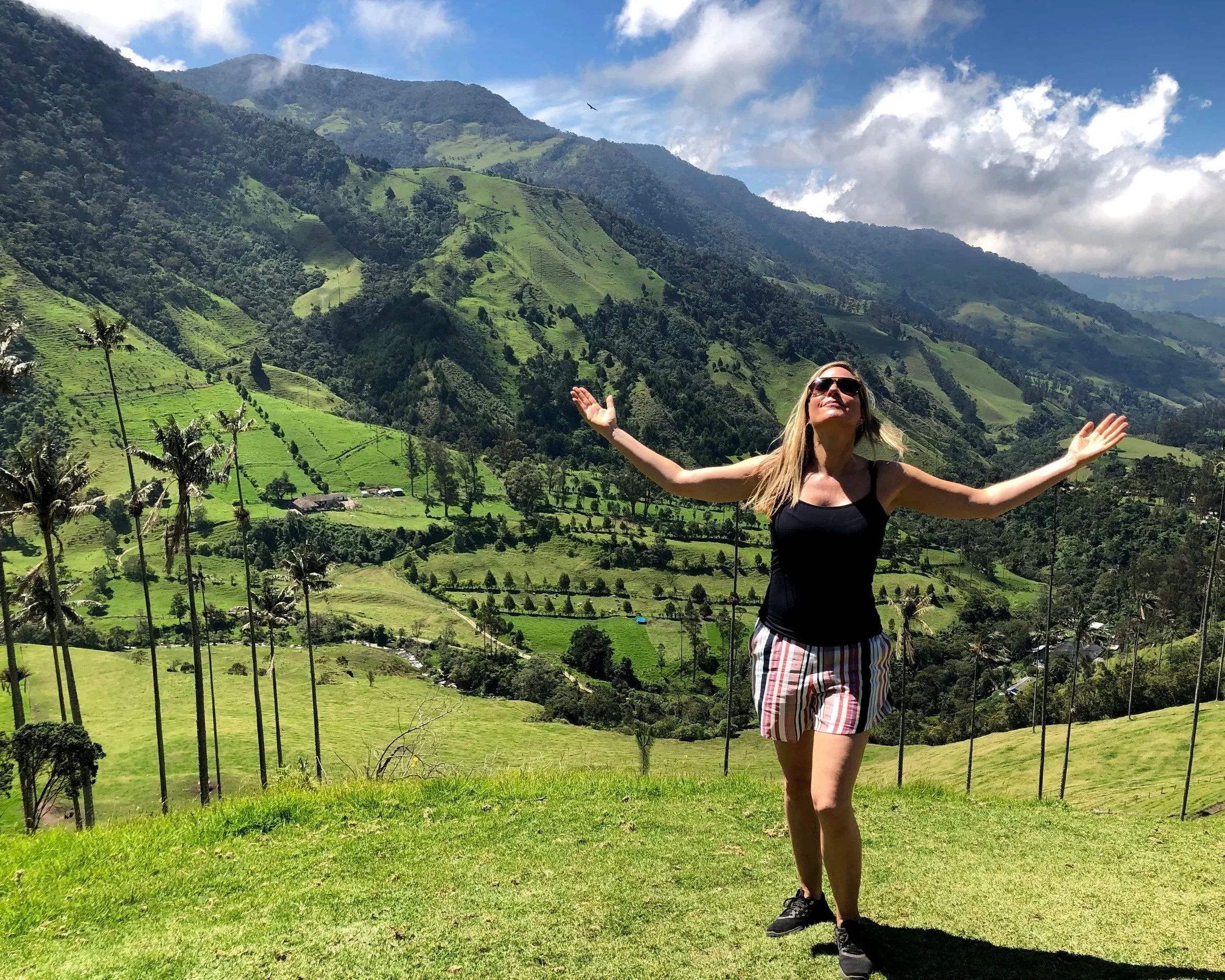 A woman in sunglasses with arms outstretched standing on a grassy hill in a lush green valley surrounded by mountains and hills, under a partly cloudy sky.