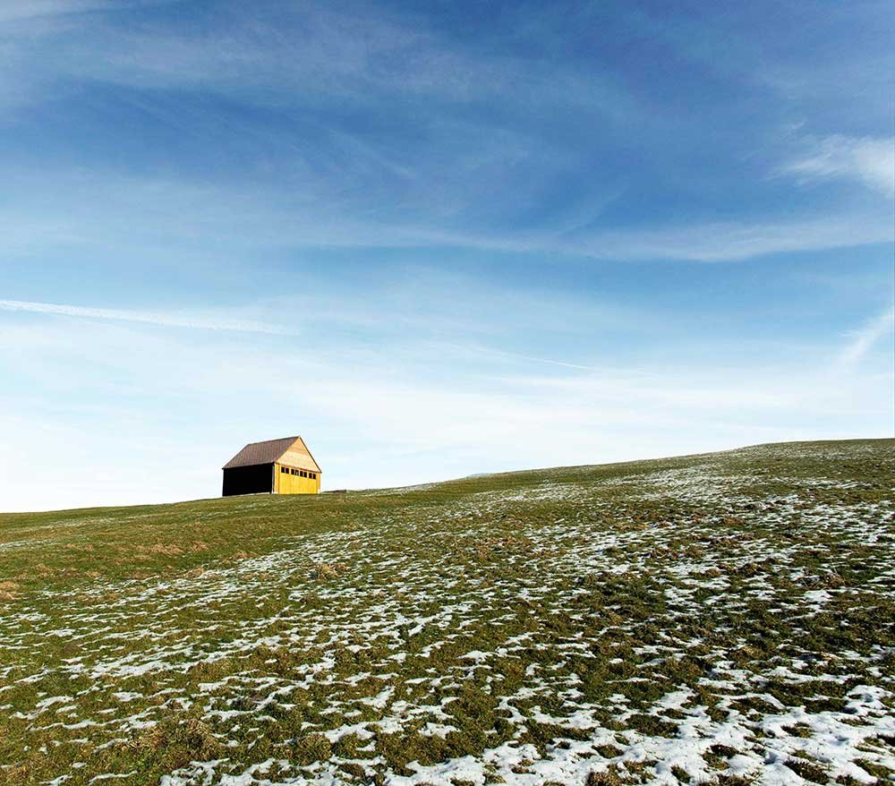 Grass hill meets a blue sky, where a yellow wooden hut sits