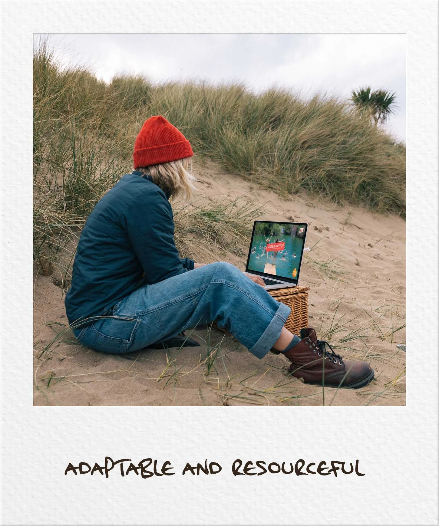 Woman wearing jeans adn red hat sitting on the sand. working on a laptop that is resting on a wicker basket