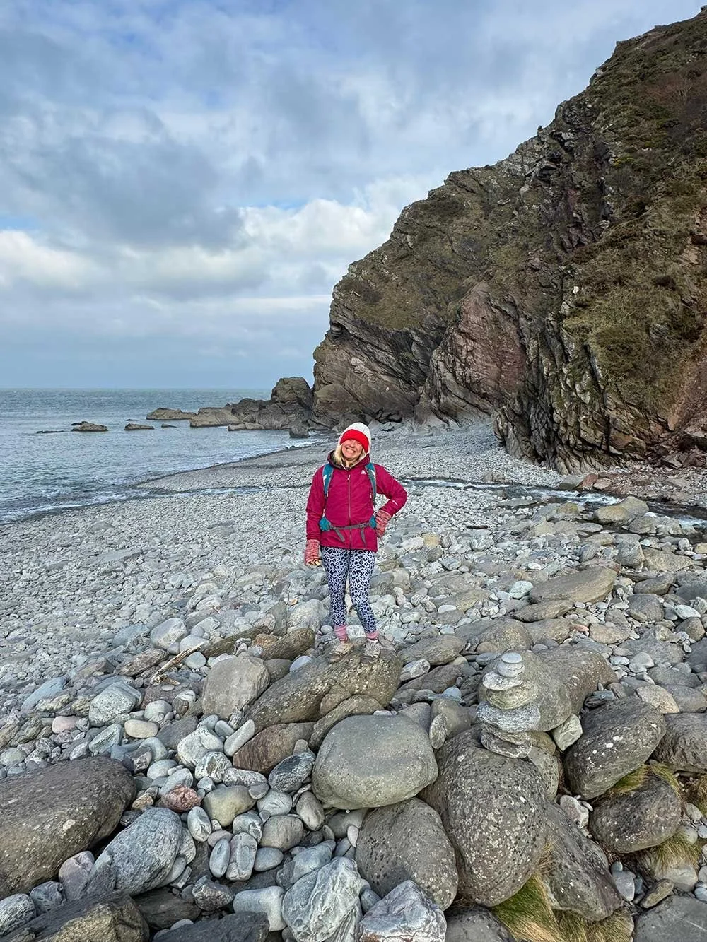 Woman on a rocky beach, wearing pink and blue hiking clothes, smiling at the camera with one hand on hip