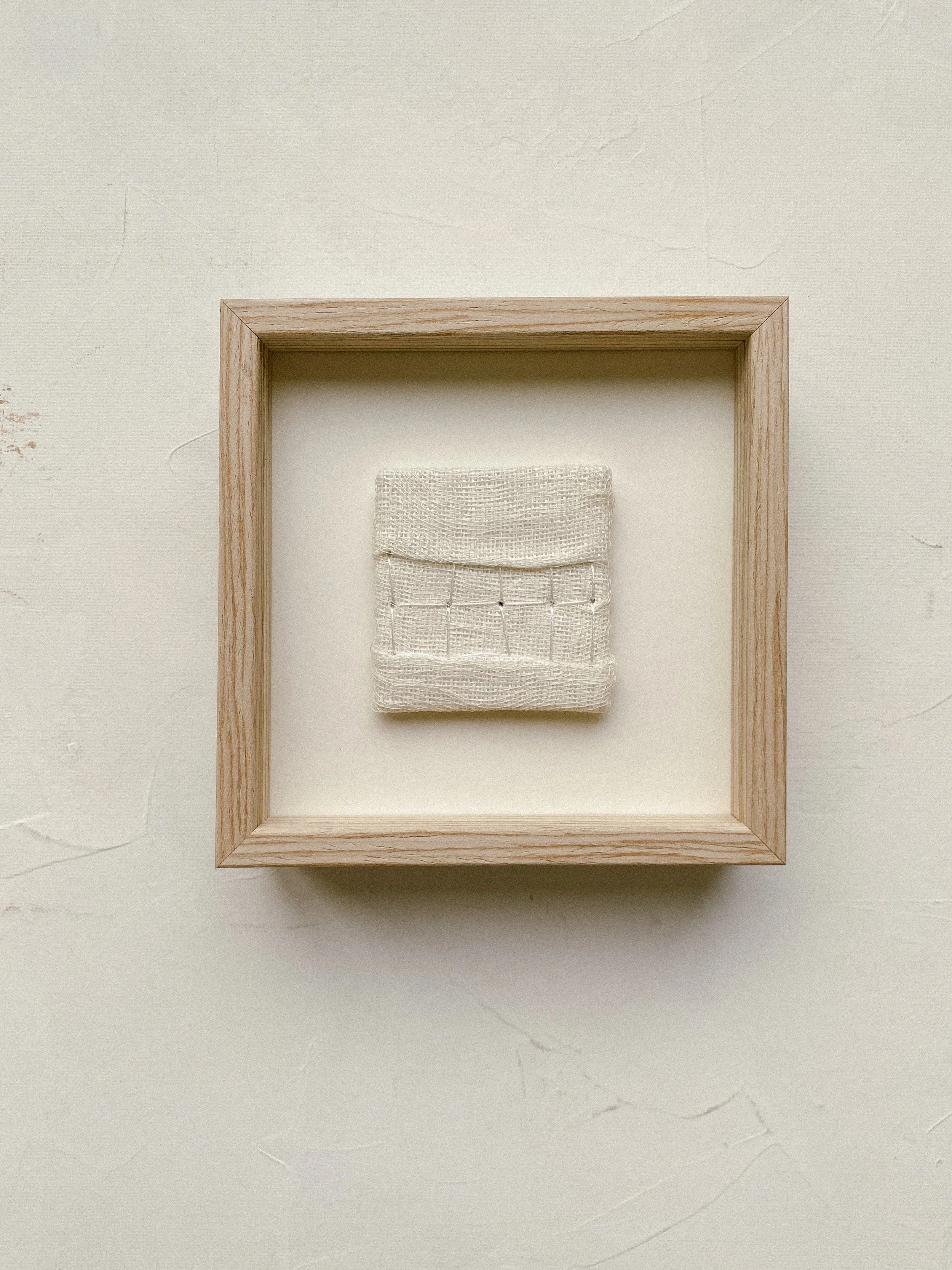 Minimal textile wall art is hanging on the wall framed in a white glazed wooden frame, dimensions are 10 by 10 centimeters.