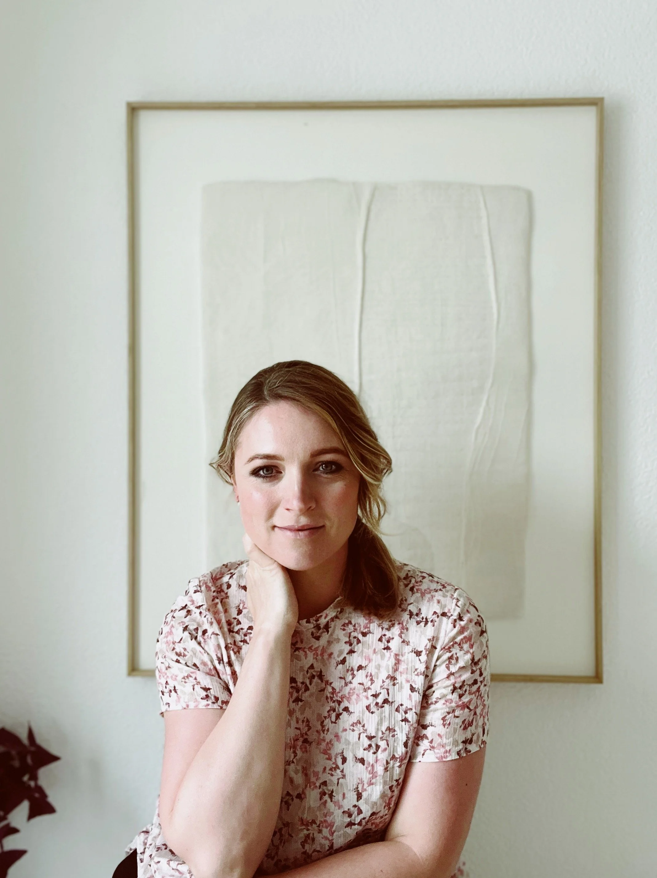 Female artist sitting in front of a hanging textured natural toned wall art piece and looking at the camera.