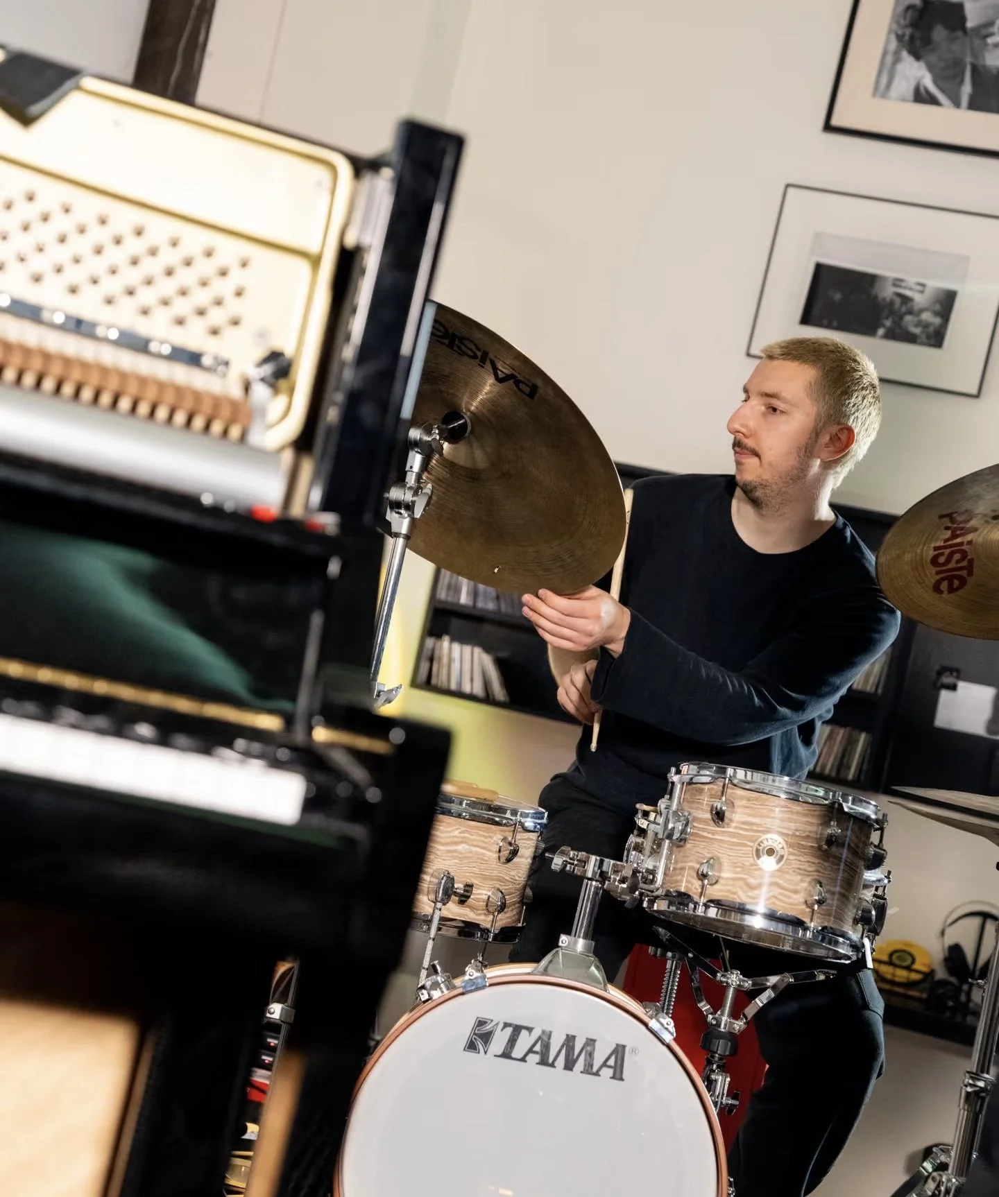 A man playing cymbals and drums in a room decorated with black and white photographs. A digital piano is in the foreground.