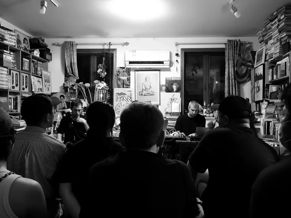 A black and white photo of a small music performance or gathering in a cozy, cluttered room with books and artwork on the walls and shelves, featuring with a group of people seated and two musicians at the front, one operating a laptop and sound equi