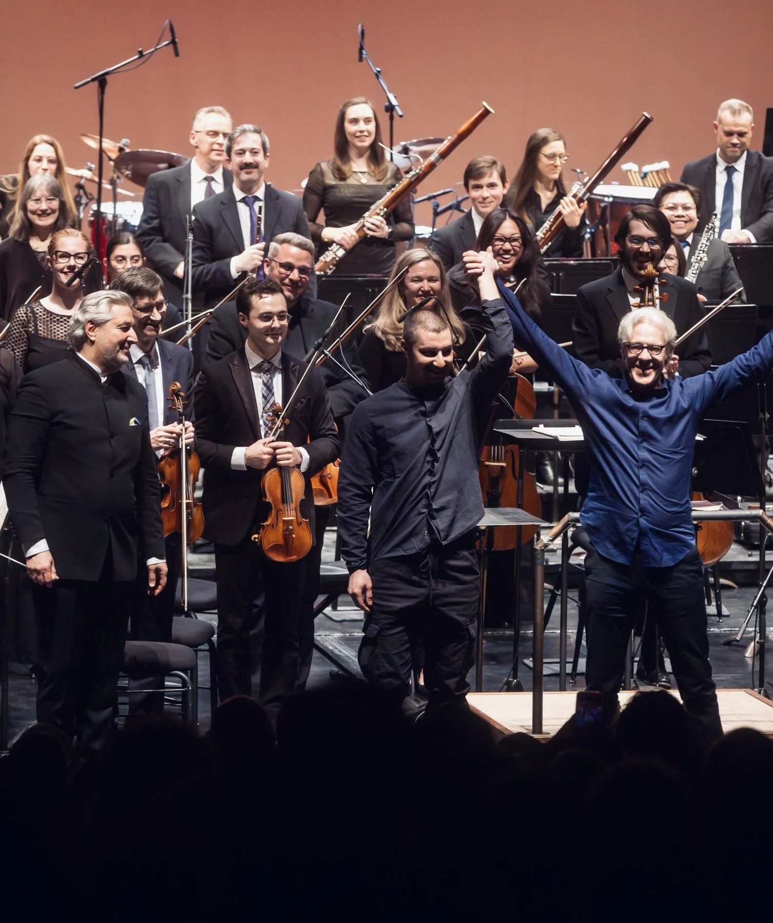 Orchestra performers on stage, some holding violins and wind instruments, celebrating with conductor. Audience at the bottom.