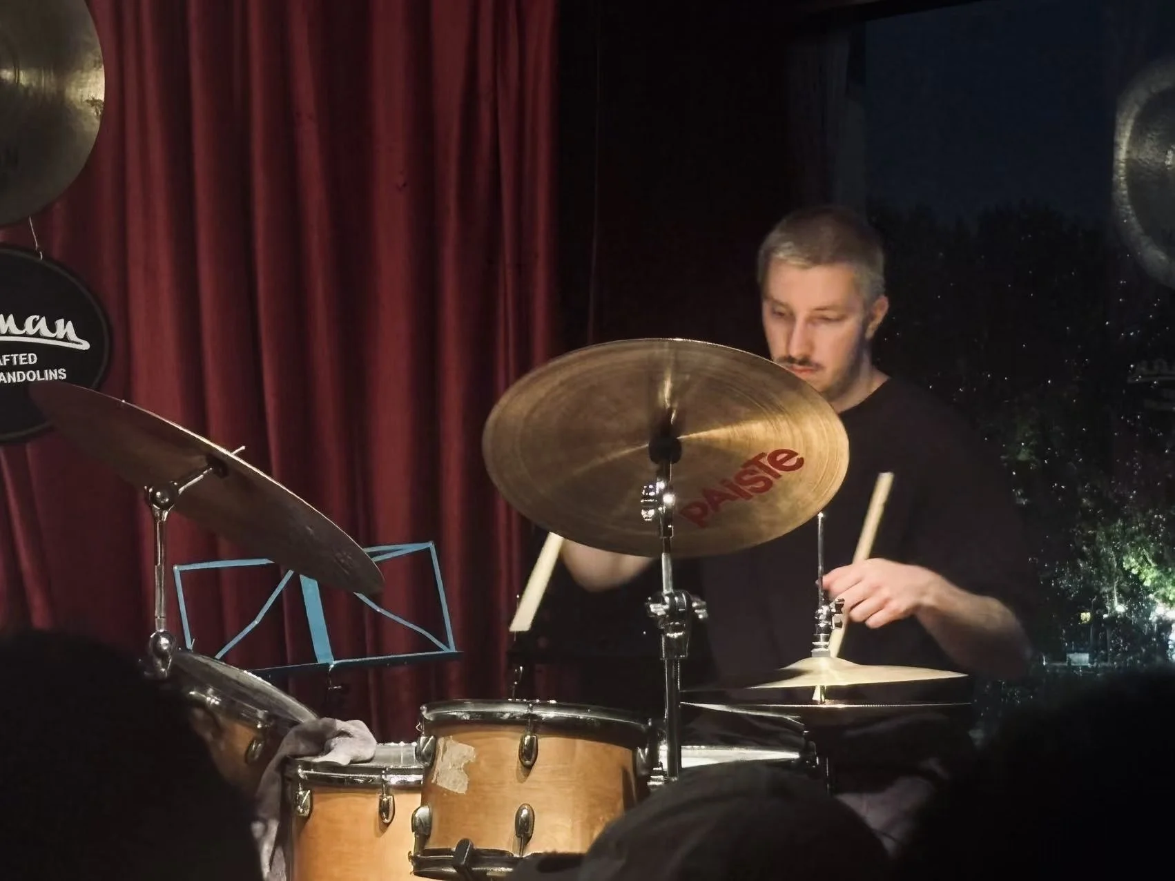 A young man with short hair playing drums on stage, with a red curtain in the background and stage lighting.