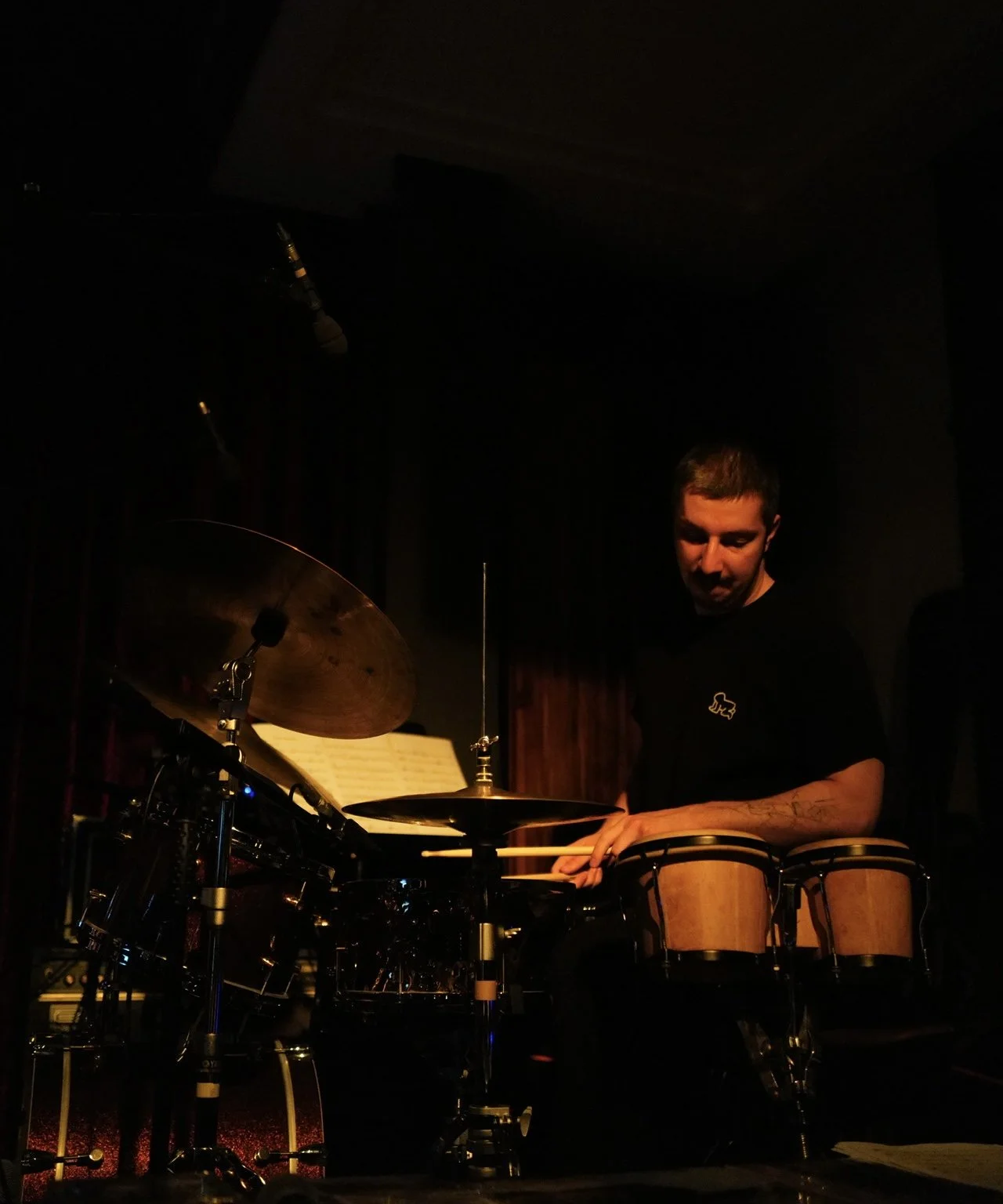 A young man plays percussion instruments on stage, including bongos, with a drum set and cymbals, in a dark venue.